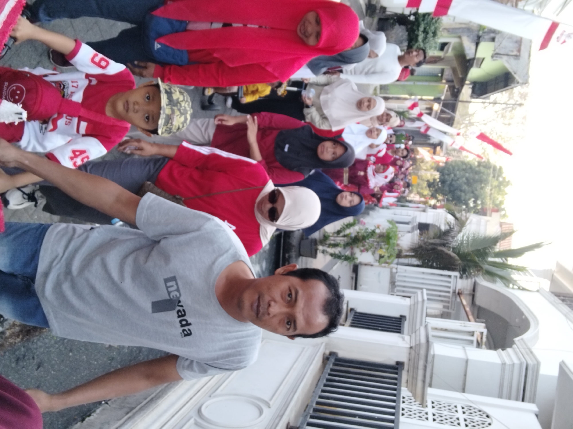 In a street scene in Berbek, Indonesia, a group of people, including men, women, and children, are participating in what appears to be a parade or procession. The participants are dressed in various colored clothing, with a notable presence of red and white attire, possibly indicating a national celebration. Some individuals are wearing headscarves, suggesting a cultural context.
In the foreground, a young boy wearing a camouflage baseball cap and a red and white t-shirt with numbered insignias is visible. He has a red backpack with a smiley face design slung over his shoulder. Beside him, a man in a grey t-shirt with the word "nevada" printed on it is walking. The man is looking directly at the camera. Behind them, other participants are moving along the street, with women in red and white outfits and headscarves, and men in white shirts.
The environment includes buildings on either side of the street, one of which is white with decorative architectural elements and a black metal fence. Indonesian flags, red and white banners, are visible strung up along the street and on buildings, further suggesting a celebratory or nationalistic event. The ground is paved with asphalt. The lighting suggests it is daytime, possibly late afternoon due to the shadows.