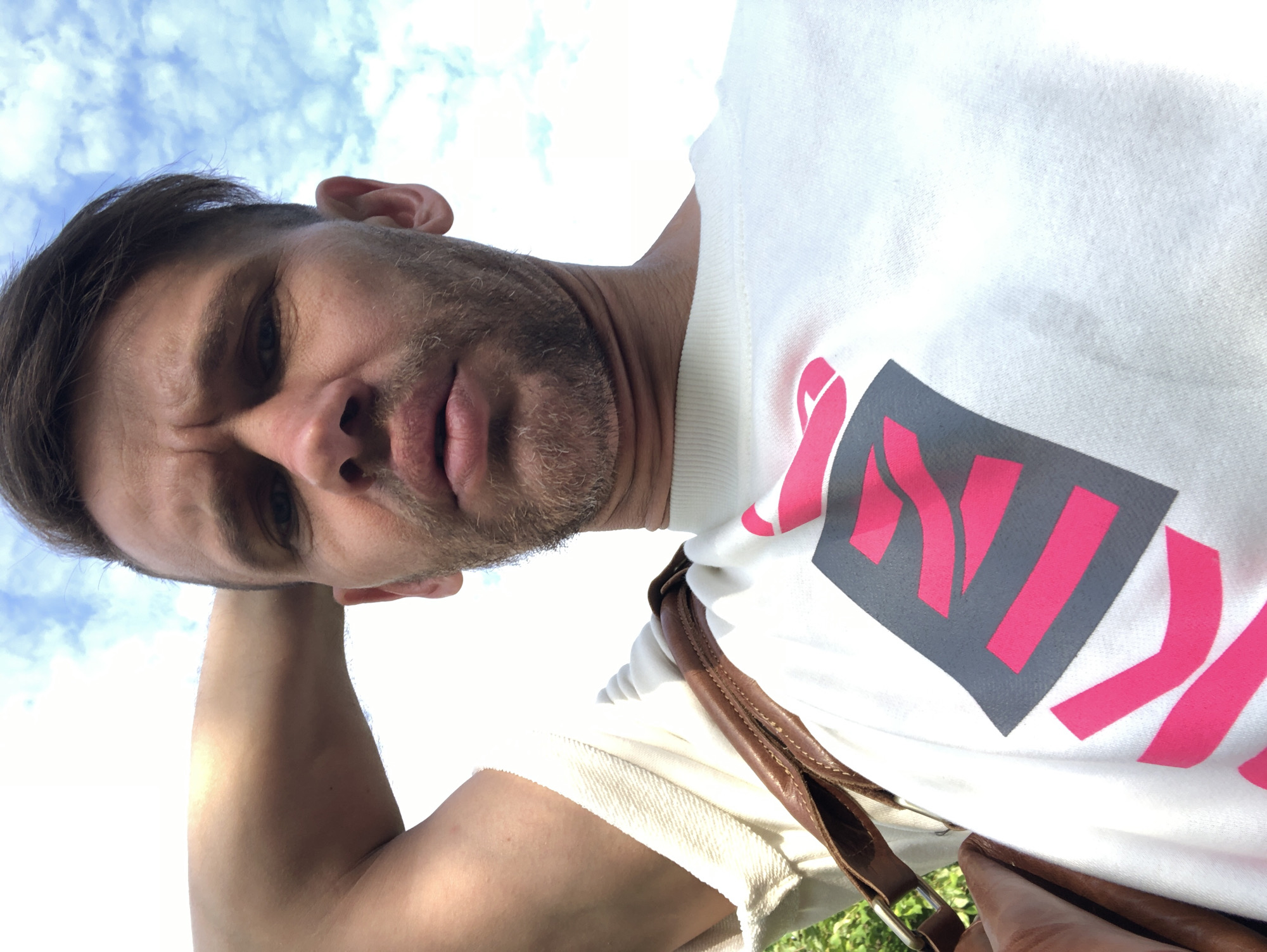 A man with short brown hair and a beard takes a selfie outdoors. He is wearing a white t-shirt with a large graphic design on it, featuring pink and gray shapes. A brown leather strap, likely from a bag, is visible across his shoulder. The background is a bright blue sky with scattered white clouds, suggesting it is daytime. The man is holding his left arm up behind his head, and his facial expression is serious. The location is not specified in the image or provided context.