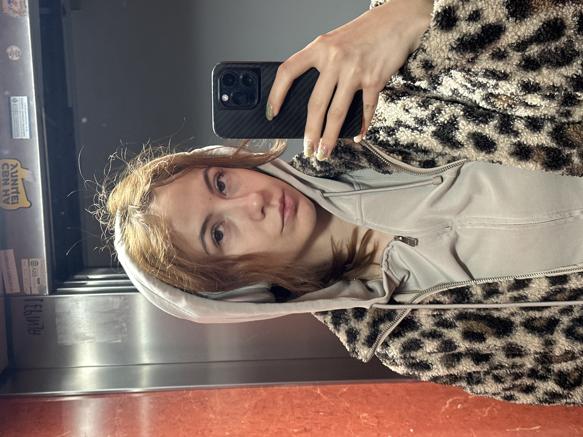 A young woman is taking a mirror selfie, captured in an indoor setting. She has light brown to auburn hair that frames her face, and she looks directly into the camera with a neutral expression. Her right hand, adorned with long nails featuring a light greenish-yellow abstract design, holds a dark-cased smartphone, likely an iPhone given the visible camera cluster. She is dressed in a light grey hooded sweatshirt layered under a fluffy leopard-print jacket.
The background reflects a metallic surface, possibly an elevator wall or a polished panel, with faint smudges and reflections. A vibrant reddish-brown floor is visible at the bottom of the frame. On the reflective surface, a sticker with the word "AUNTIE!" is visible in reverse, alongside other partial, reversed text that appears to be "AH N8Ɔ HV". Lower down on the metallic panel, the faint, reversed text "ALINJ3" can be discerned. The image is taken indoors, with no specific time of day or weather cues other than the artificial lighting.