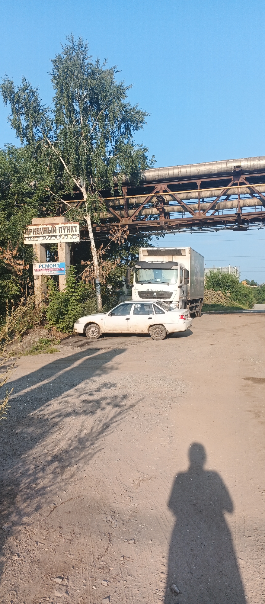 A white sedan is parked on a dirt road next to a large white truck. The sedan is facing left and the truck is behind it, facing right. Above the vehicles, a large industrial pipe structure spans across the frame, supported by metal beams and concrete pillars. A white sign with Russian text is visible on one of the pillars, and another smaller blue sign with "РЕМОНТ" (Repair) is also attached to a pillar. A birch tree with green foliage stands to the left of the signs. The sky is clear and blue. The scene appears to be in an industrial area.