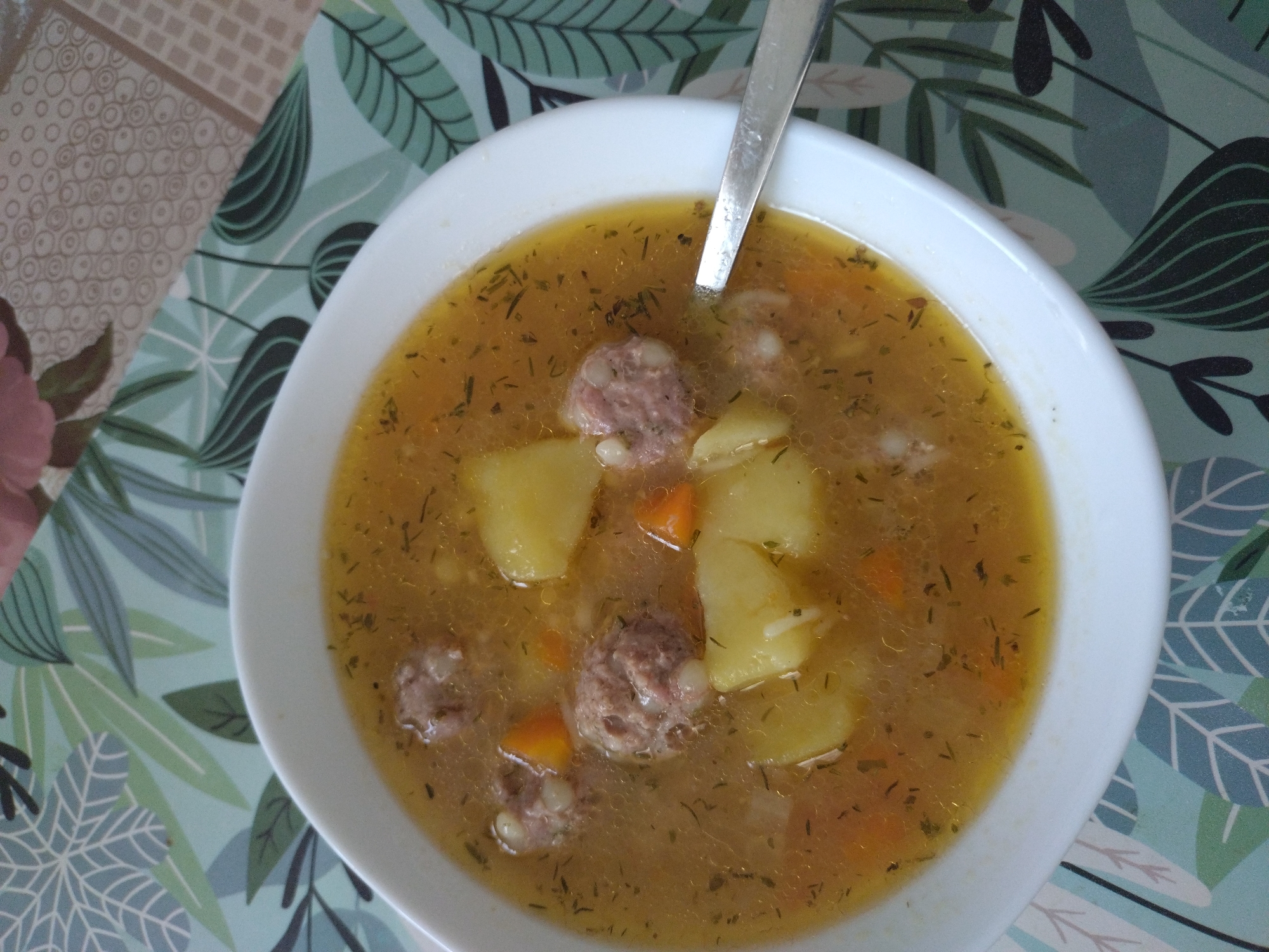 A close-up shot depicts a bowl of soup on a patterned surface. The soup appears to contain meatballs, potatoes, and carrots, with dill floating in the broth. A silver spoon rests in the soup. The background consists of a table covering with a floral and geometric pattern. The image was likely taken indoors, and the location in the city of Stara Zagora, Bulgaria is not discernible from the visual information provided.