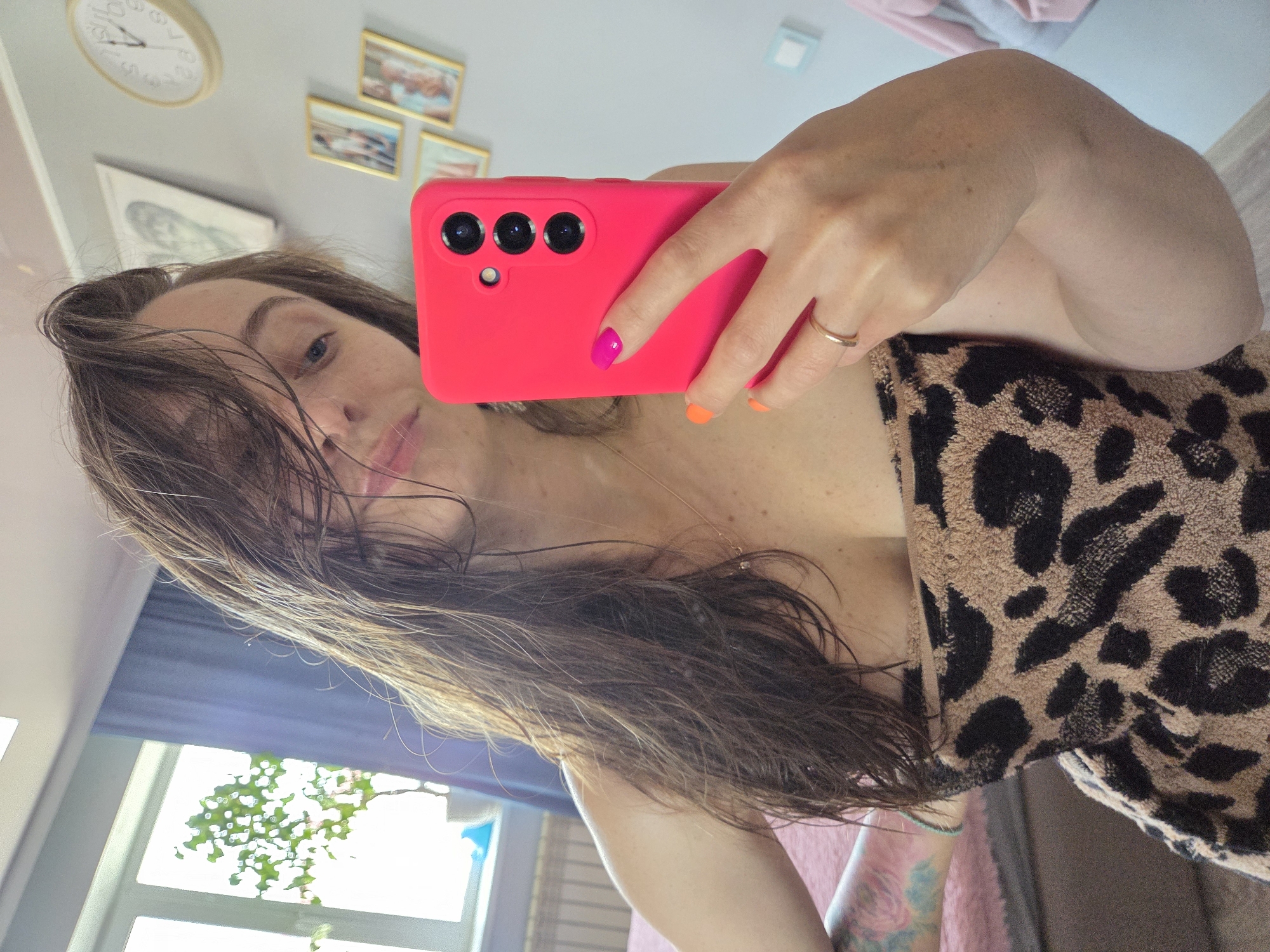 A woman is taking a selfie in an indoor setting. She is holding a bright pink smartphone with three camera lenses. Her wet, shoulder-length brown hair is partially obscuring her face. The woman is wearing a ring on her finger. She is wrapped in a tan and black leopard-print towel. A window is visible in the background, showing green foliage outside. A clock and framed pictures hang on the wall. The location in Birobidzhan, Russia is not discernible from the image.