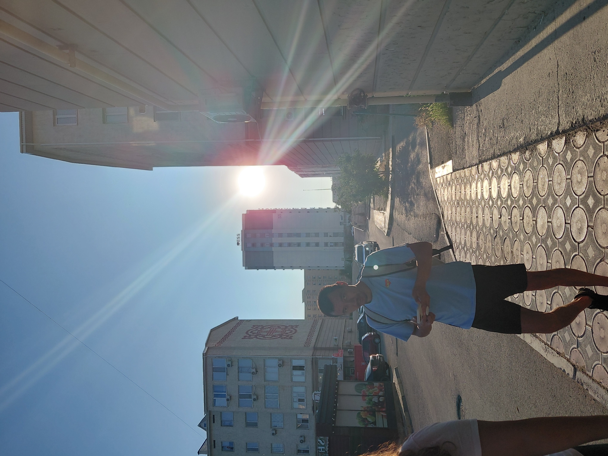 In Aktau, Kazakhstan, under a bright, sun-drenched sky, a young man stands on a sidewalk paved with decorative circular tiles. He is dressed in a light blue t-shirt and black shorts, and wears a backpack. He is holding a smartphone in his hands and appears to be looking at it. Behind him, the street is lined with various buildings, including a multi-story residential building with a large, decorative emblem on its facade, and a taller, modern building with the "ESS" logo visible. Several cars are parked along the street and a few are in motion. The sun is low in the sky, casting long shadows and creating lens flare effects across the scene. The overall impression is of a warm, sunny day in a city environment.