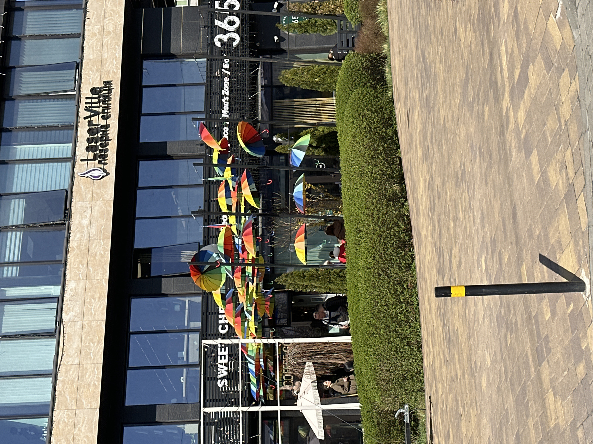 The photograph depicts a commercial building in Kyiv, Ukraine, likely a shopping center or business complex. The building features large glass windows and a tan facade with the signage "LaserVille Laser & Aesthetic Clinique." In front of the building, a decorative display of colorful umbrellas hangs above the entrance. Several shops are visible, including "Sweet Chef" and "Coffee Go." People are seen entering and exiting the building. A meticulously maintained hedge separates the building from a paved walkway. A black pole with a yellow band is attached to the ground. The building also has a sign that reads "Men's Zone" and the number "365".