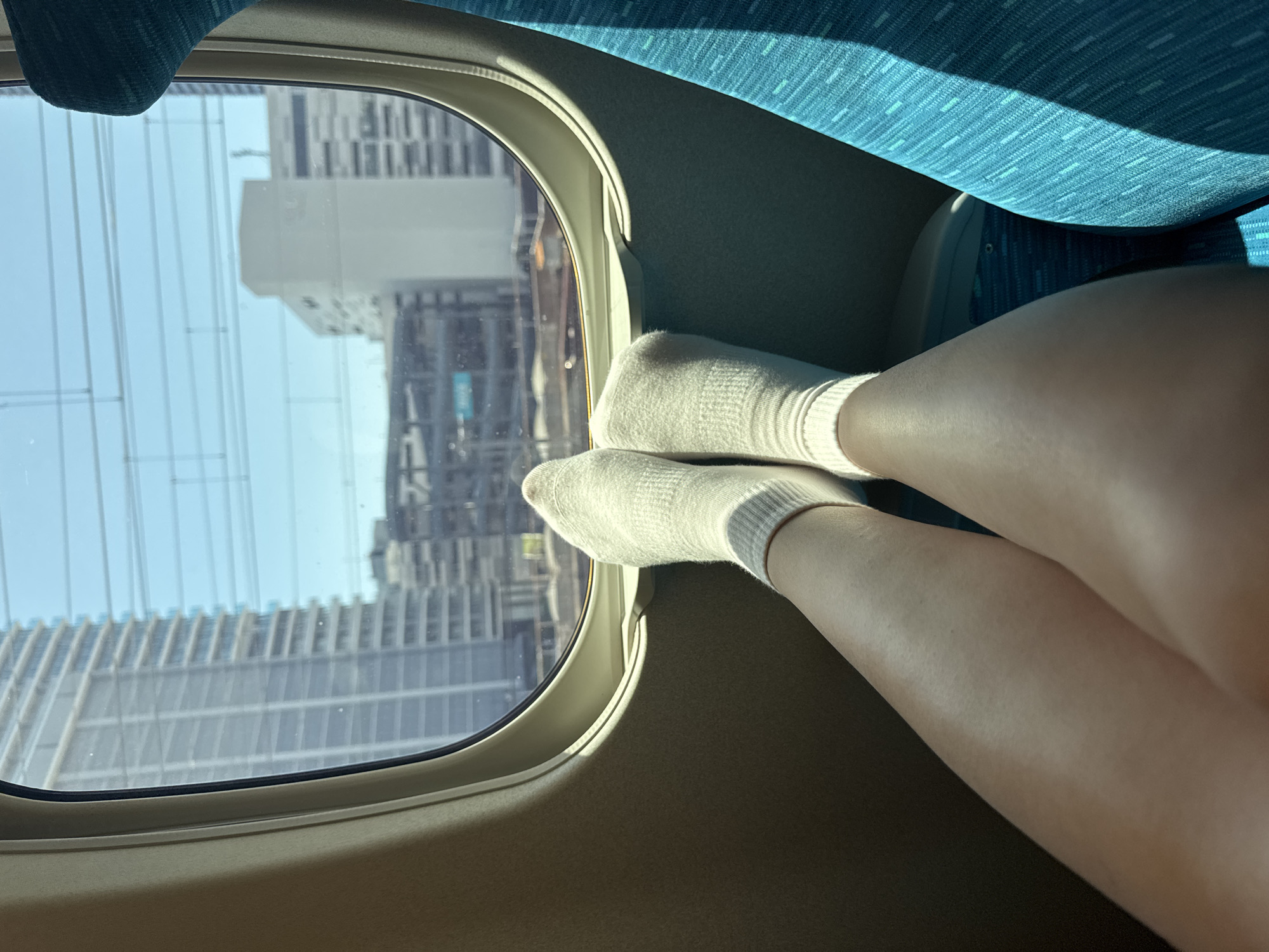 From the interior of a train car, a view of a city scene through a window is presented. The feet, clad in white socks, are placed against the bottom edge of the window. Outside, buildings line the landscape. Based on the urban architecture and transportation system, this image is likely taken in Hiroshima, Japan. The scene suggests a casual moment during a train journey.