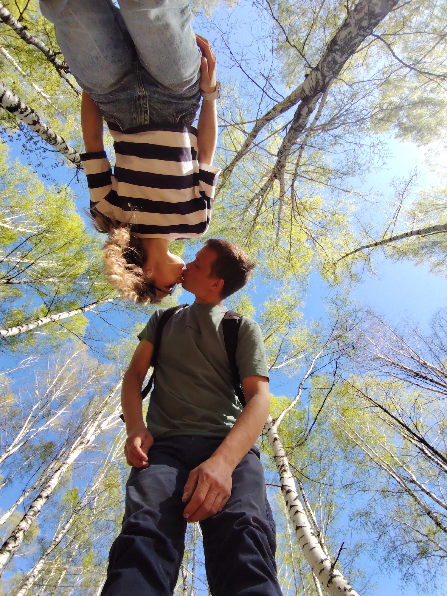 In Chaikovsky, Russia, a man and a woman are kissing in a birch forest. The woman is upside down, suspended from the branches of a tree, wearing a striped shirt and jeans. The man is standing on the ground, facing the woman, wearing a green shirt and dark pants with a backpack. The sky is visible through the trees, and the leaves are a vibrant green.
