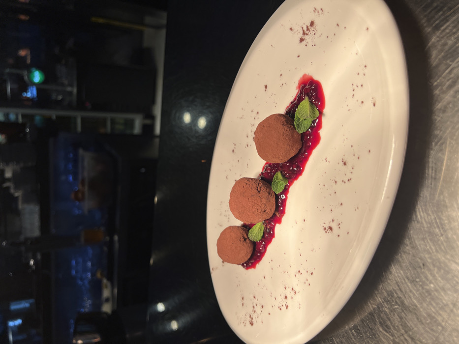 A beautifully plated dessert is the central focus of this image, presented on a long, oval white plate. Three round chocolate truffles are arranged diagonally across the plate. Each truffle is generously dusted with cocoa powder, giving them a rich, matte brown appearance.
Beneath and around the truffles is a vibrant, deep red coulis or berry sauce, which has been artfully swirled or spread in a wavy line. Small, fresh mint leaves are strategically placed among the truffles and the red sauce, adding a touch of green contrast to the dark chocolate and red fruit. The plate itself is glossy and pristine, with subtle speckles of cocoa powder scattered around the dessert, enhancing the presentation.
The background on the left side of the image is dark and out of focus, suggesting an indoor setting with low lighting, possibly a restaurant or bar. Blurred lights and reflections are visible, including a distinct green light towards the upper left. This blurry background emphasizes the dessert in the foreground. The surface beneath the plate on the far right appears to be a dark, possibly metallic or brushed steel countertop or table, reflecting some light sources. The overall impression is one of an elegant, well-prepared dessert served in a refined evening environment. There is no visible text in the image.