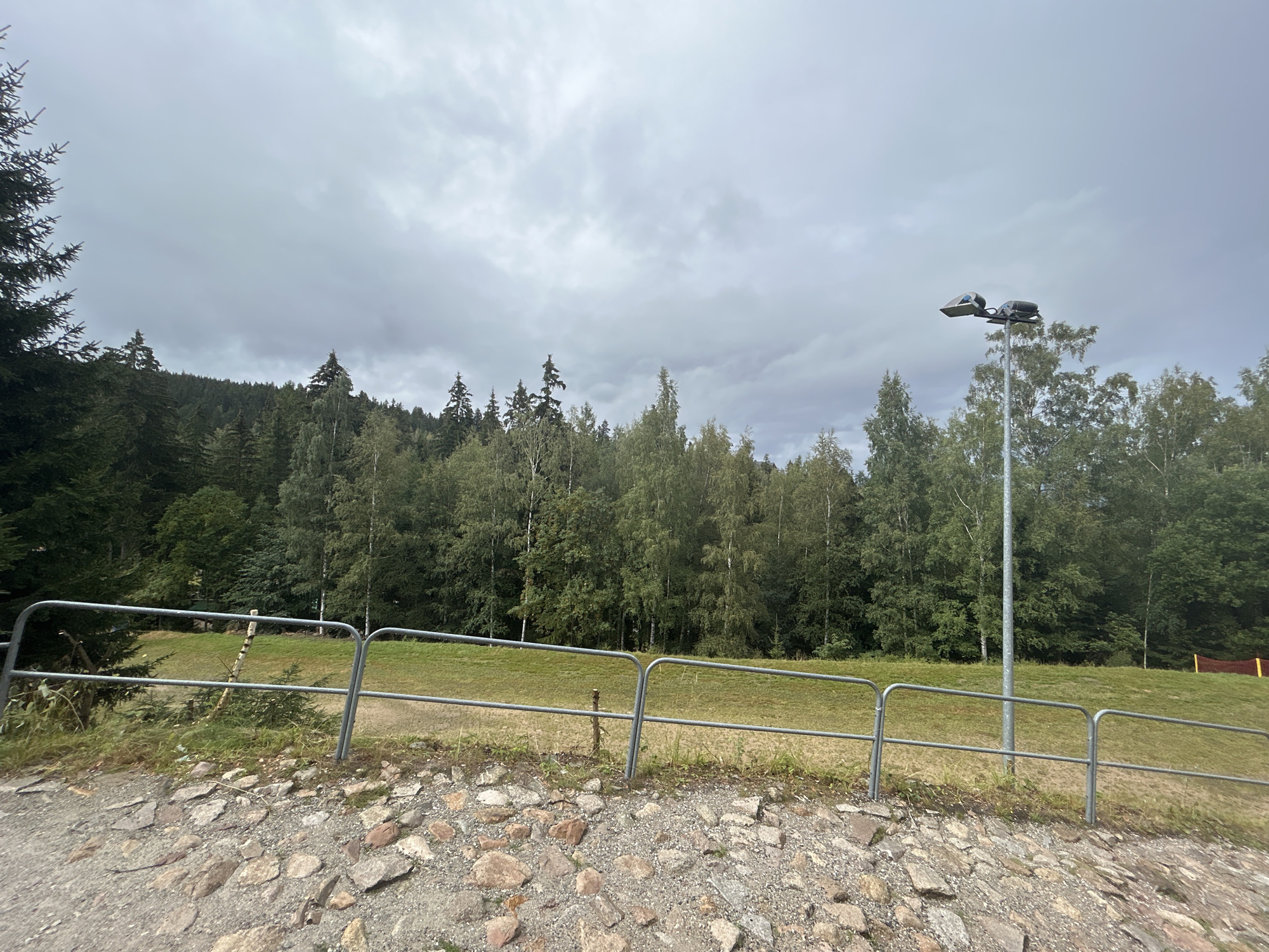 The image depicts an outdoor scene with a cloudy sky. In the foreground, a pathway is covered with rocks and gravel. A metal railing runs horizontally across the middle ground, separating the pathway from a grassy area beyond. A tall metal lamppost with two light fixtures is visible on the right side of the image, positioned behind the railing. A dense forest borders the grassy area, extending into the background. The overall environment suggests a park or recreational area. This scene is located in Karpacz, Poland.