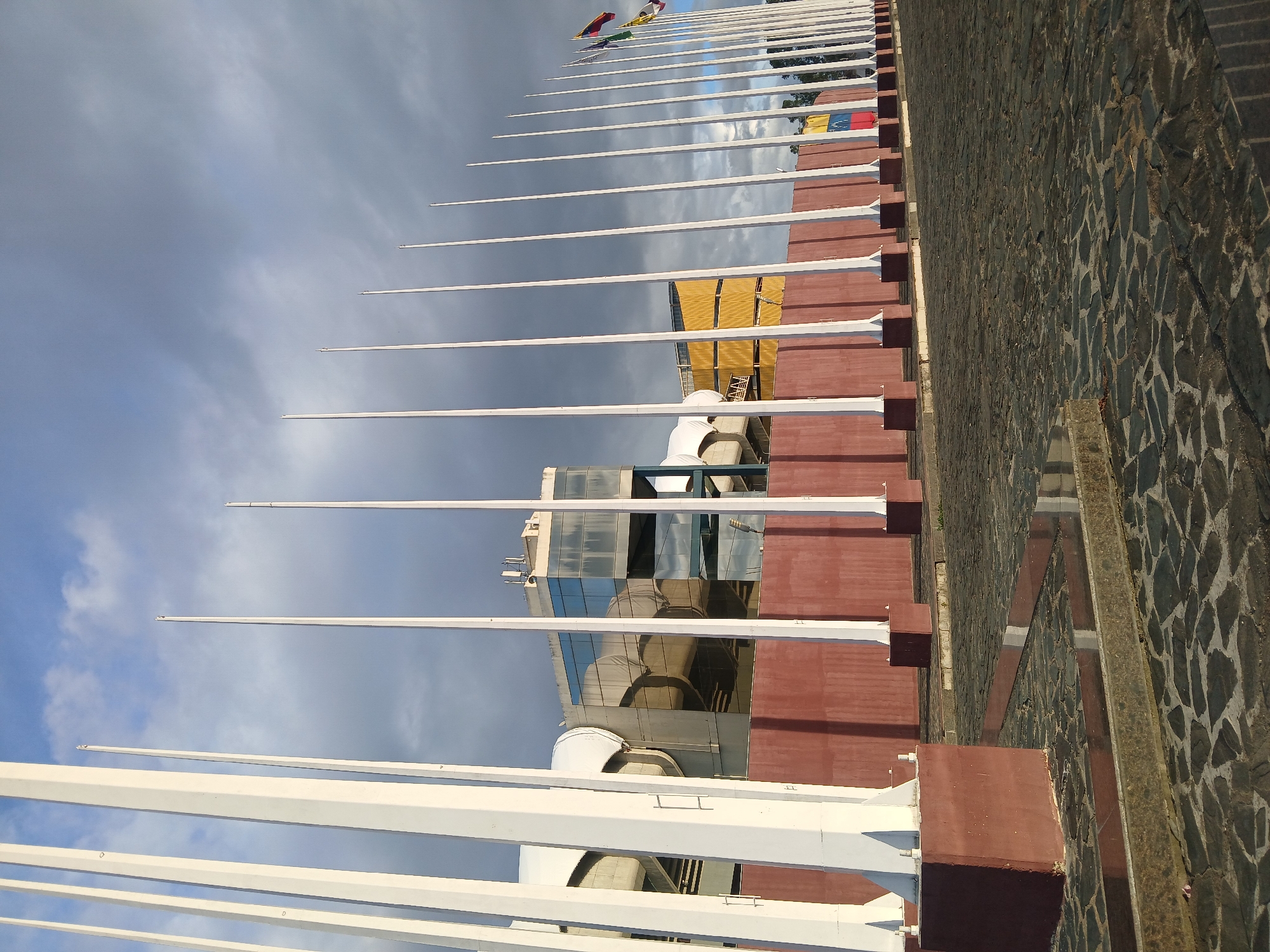 The image shows a row of flagpoles without flags, except for a few at the distant end, in Ciudad Guayana, Venezuela. The flagpoles are white and are anchored to red-brown rectangular bases. A building with reflective glass panels and white rooftop equipment is visible in the background. The sky is cloudy with shades of blue and gray. A stone-paved surface runs parallel to the flagpoles on the right side of the image. The image is taken from a low angle, looking upwards along the line of flagpoles.