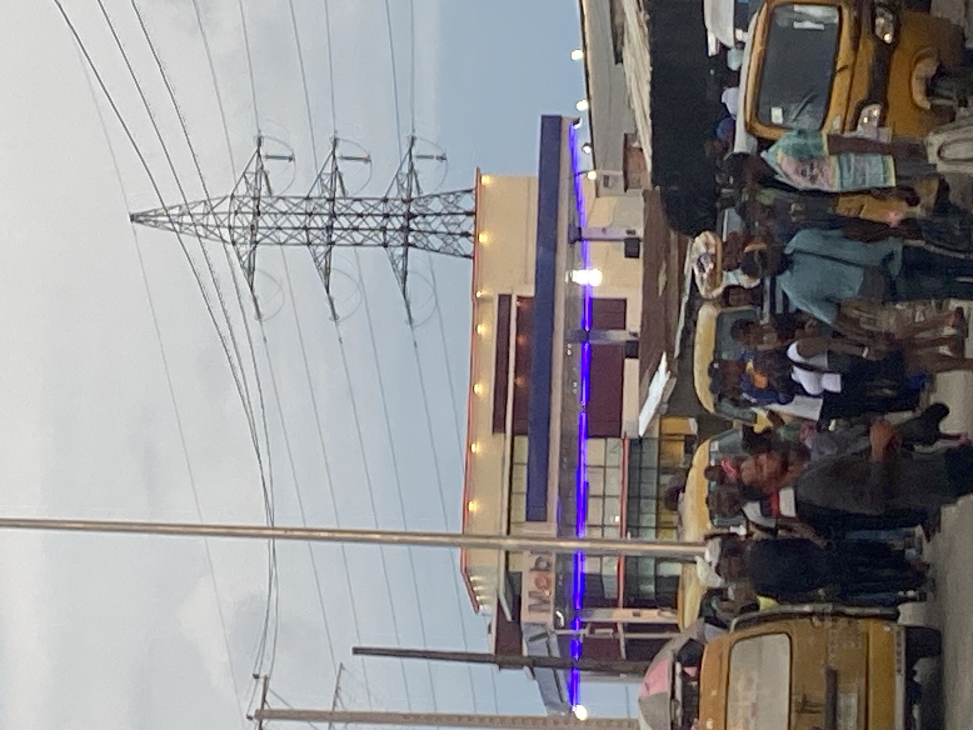 In Lagos, Nigeria, under a cloudy sky, a bustling street scene unfolds. A tall, lattice metal power tower dominates the background, supporting multiple high-voltage power lines that stretch across the frame. Below it, a two-story building, possibly a gas station given the visible "Mobi" signage on the facade, features a modern design with a cream-colored upper level adorned with yellow accent lights and a lower level with blue neon lighting.
The street is crowded with people, many appearing to be commuters or pedestrians. Yellow minibusses, a common mode of public transport in Lagos, are parked or in motion, with one yellow minibus prominently positioned on the left side of the image. A yellow auto-rickshaw, also known as a "keke," is visible on the right. People are engaged in various activities, including walking, standing, and carrying items, suggesting a busy urban environment. The overall atmosphere is one of daily life in a densely populated city.