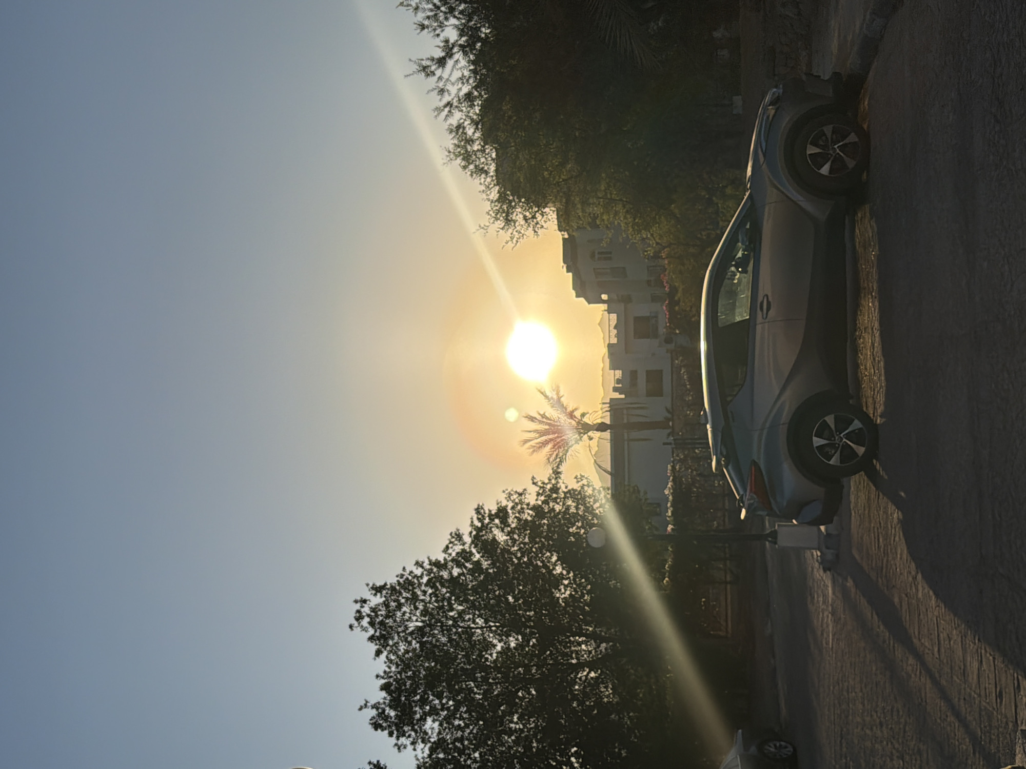 A silver sedan is parked on a paved surface, casting a long shadow in the warm glow of the setting sun. The car is positioned horizontally in the frame, facing right. In the background, a modern building with multiple windows is visible, partially obscured by lush green trees. A palm tree stands out against the sky, its fronds silhouetted by the sun. The sky is a clear, soft blue, transitioning to a warm yellow near the horizon where the sun is setting. The overall scene evokes a sense of tranquility and the peaceful end of a day, likely in a warm, possibly tropical or sub-tropical, location like Sharm El Sheikh, Egypt. There is no visible text in the image.