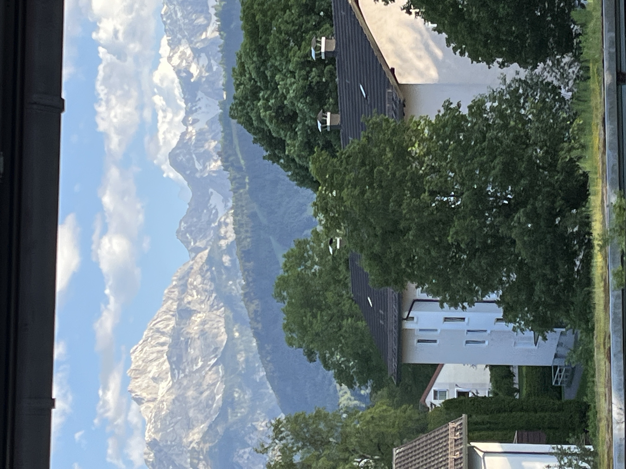 The image presents a view from a window in Garmisch-Partenkirchen, Germany. Visible is a scenic mountain range with rocky peaks and some snow cover against a backdrop of a partly cloudy blue sky. In the mid-ground, there are buildings, specifically houses with dark tiled roofs and white walls, partially obscured by green trees. The immediate foreground includes the frame of the window from which the picture is taken. The composition is predominantly vertical, emphasizing the height of the mountains and the buildings. There are no people visible.