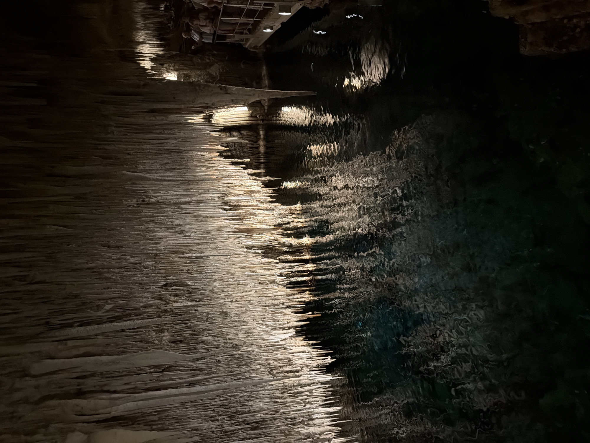 The image shows the Caves of Drach in Manacor, Spain. The left side of the image depicts a rock formation with stalactites. The right side of the image depicts water. The stalactites are white and are illuminated by artificial light. The water is dark and reflects the light.
