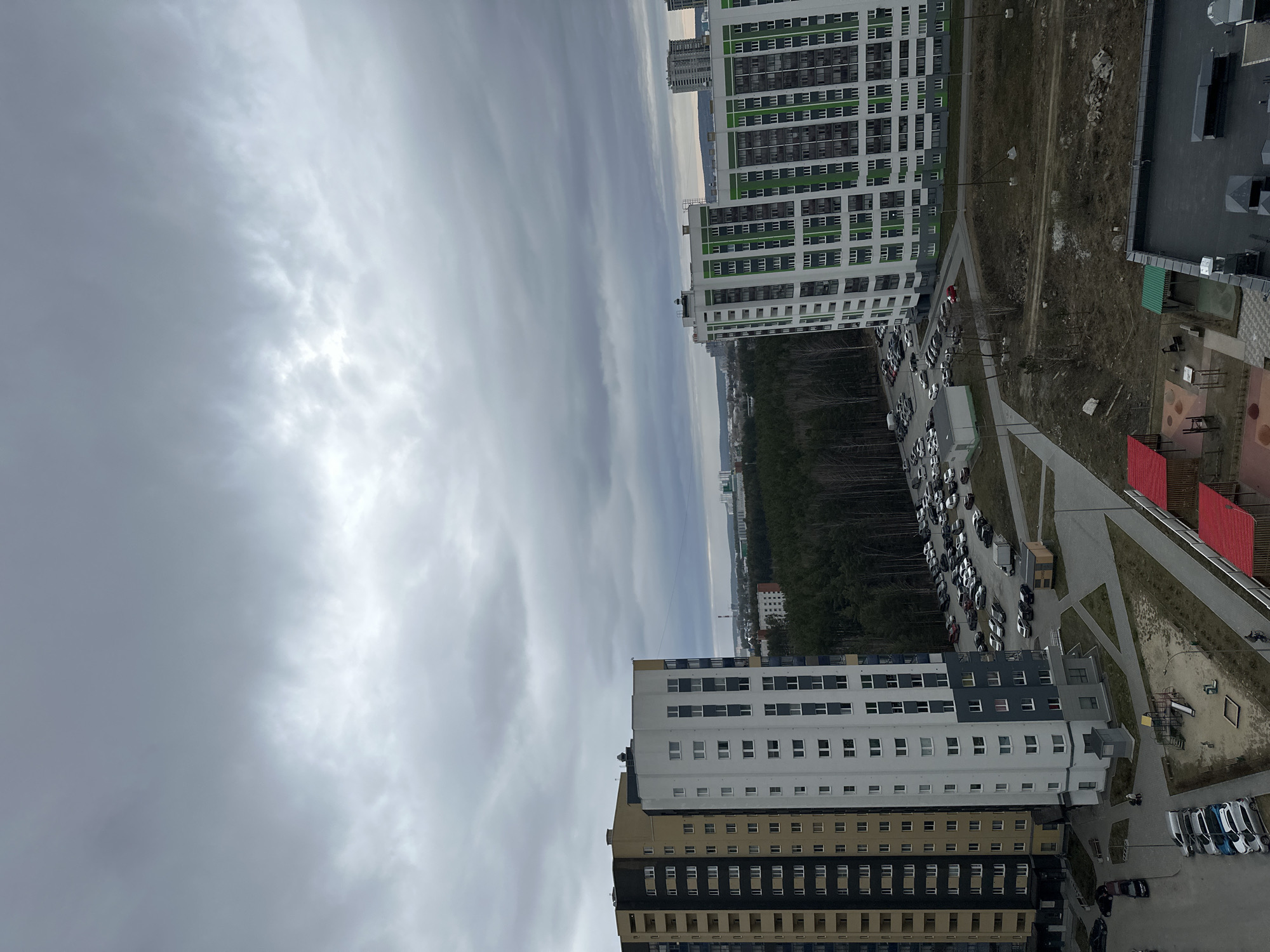 The image shows an urban scene in Yekaterinburg, Russia, taken from a high vantage point. The sky is overcast with striated cloud formations. Two high-rise apartment buildings are prominent in the frame. One building has a white and green facade, while the other has a more complex color scheme with black, yellow, and white sections. A parking area filled with cars is located between the buildings and a densely wooded area. A road runs alongside the wooded area. Playgrounds and other outdoor infrastructure elements are visible.