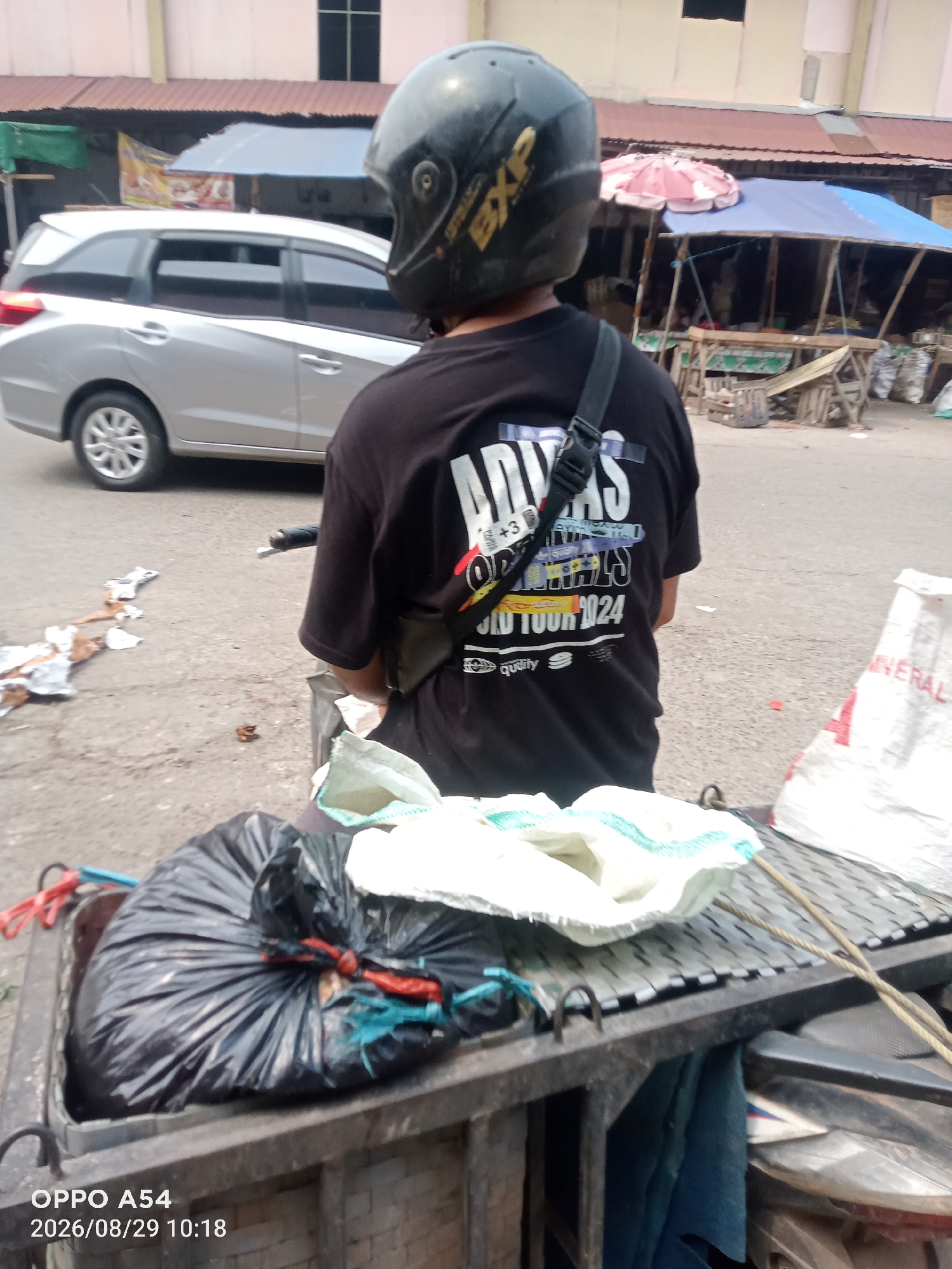 A person wearing a black t-shirt with a graphic design and a black helmet is seated on a motorbike, viewed from behind. The t-shirt has "ADIDAS" and "WORLD TOUR 1924" printed on it. The motorbike is carrying a load of trash in a black plastic bag tied with red and blue string, and two white sacks in a grey bin. In the background, a silver car is parked next to a market stall with a blue and red umbrella. The scene appears to be outdoors on a paved surface, possibly a street or marketplace. The timestamp "2026/08/29 10:18" and "OPPO A54" are visible in the lower left corner of the image. The location is identified as Jambi City, Indonesia, suggesting a typical urban environment in that region.