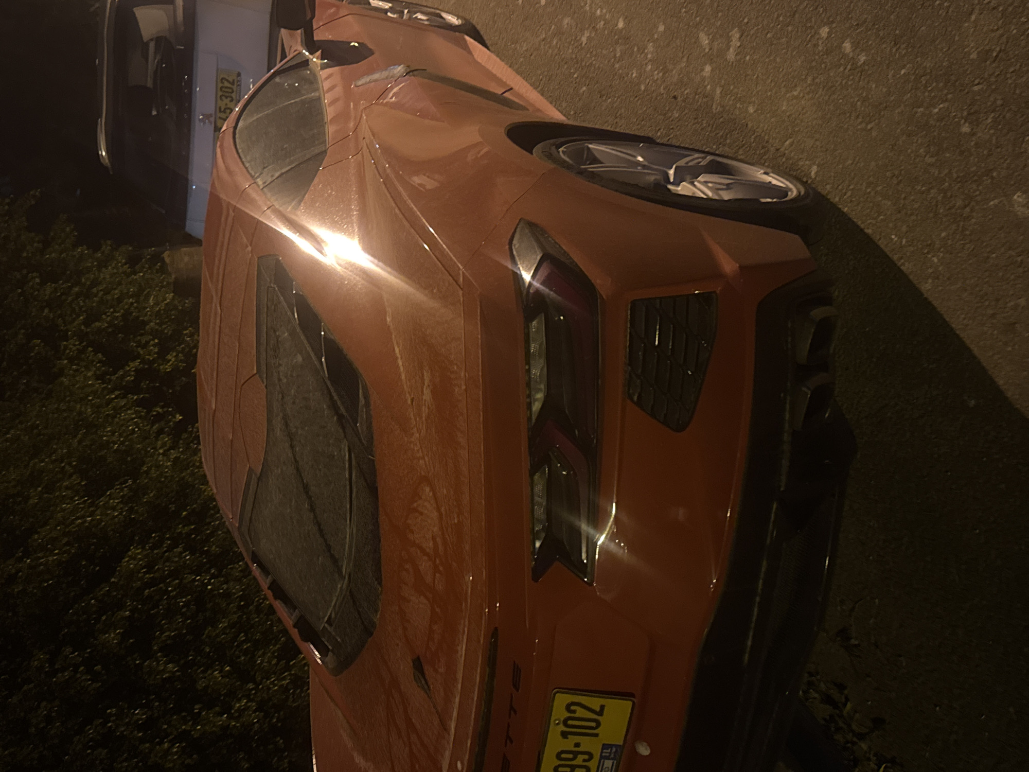 The image shows an orange Corvette parked on a street in Sdot Dan Regional Council, Israel. The license plate is partially visible. To the left, there's another vehicle and some greenery. The road surface is dark, suggesting it might be night. The car appears to be slightly dirty or wet.