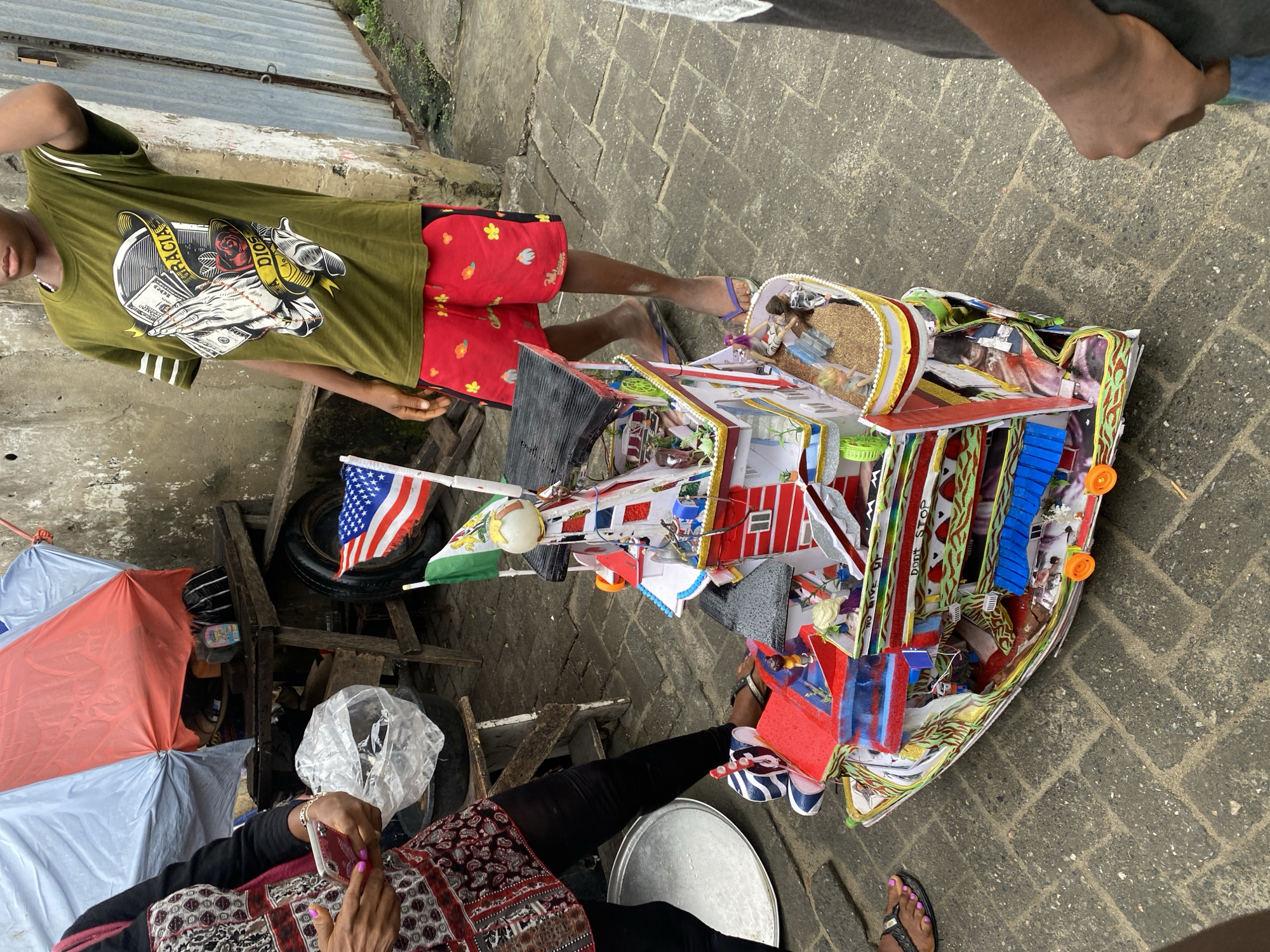A child stands next to a detailed model of a boat, likely at a street market or outdoor gathering. The boat is elaborately decorated with flags, miniature figures, and colorful patterns. The child is wearing a green t-shirt with a graphic design and red shorts. To the left, an adult is partially visible, holding a smartphone. The ground is paved with brick-like stones, and the background features a weathered wall and some market stall elements, including umbrellas and wooden structures. The scene appears to be outdoors, possibly in a town or city area.