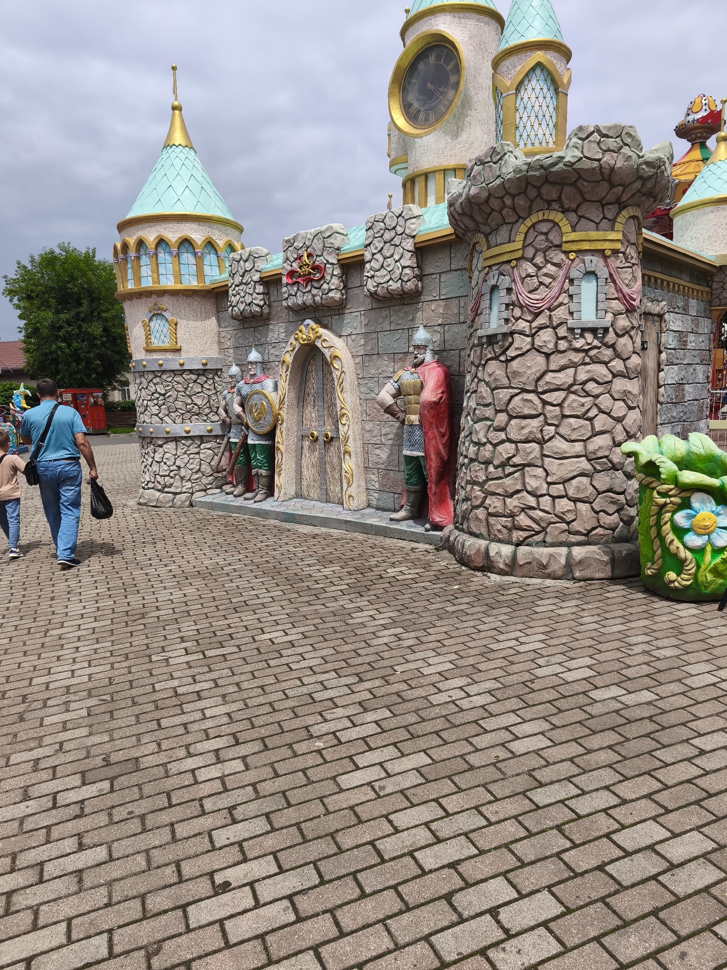 The image depicts a section of a fantasy-themed amusement park or attraction, resembling a medieval castle with exaggerated architectural features. The castle has multiple towers with turquoise, conical roofs topped with gold spires. One prominent tower features a large, circular clock face with Roman numerals. The castle walls are constructed with textured, stone-like material, adorned with gold trim and decorative elements like draped fabric motifs.
Several life-sized statues of knights in armor stand guard outside the castle entrance, one of whom is red-caped and has a hand on his hip, while others are positioned flanking the main doorway. The doorway itself is an ornate, arched entrance with a weathered wooden door and gold detailing. To the right of the entrance, a large, cartoonish planter shaped like a green leaf holds a smiling flower with a blue face and yellow center.
In the foreground, a man and a child are walking away from the castle, along a patterned brick pathway. The man, wearing a blue t-shirt and jeans, carries a black plastic bag in his right hand. The child, wearing a light-colored t-shirt and jeans, walks slightly ahead of him. The pathway is made of rectangular bricks laid in a herringbone pattern.
The background shows some greenery, including a large tree on the left, and hints of other amusement park structures, such as colorful rides and buildings with similar whimsical architectural styles. The sky is overcast with gray clouds.
The image does not contain any specific visual cues to identify the location as Vladivostok, Russia.