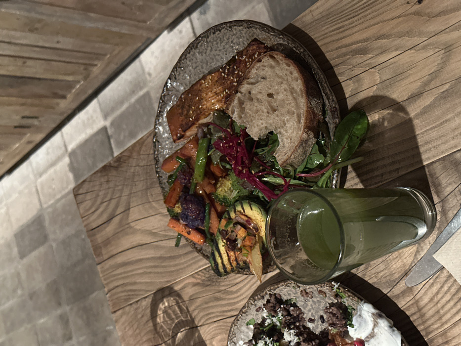 A plate presents a piece of glazed fish, sliced bread, grilled vegetables, and leafy greens. A glass filled with a green beverage sits beside the plate. Another plate is partially visible, featuring a dish of finely chopped ingredients, likely savory with a topping of white sauce or yogurt. All items are set on a wooden surface with a utensil nearby. A brick surface is visible to the left of the plates.