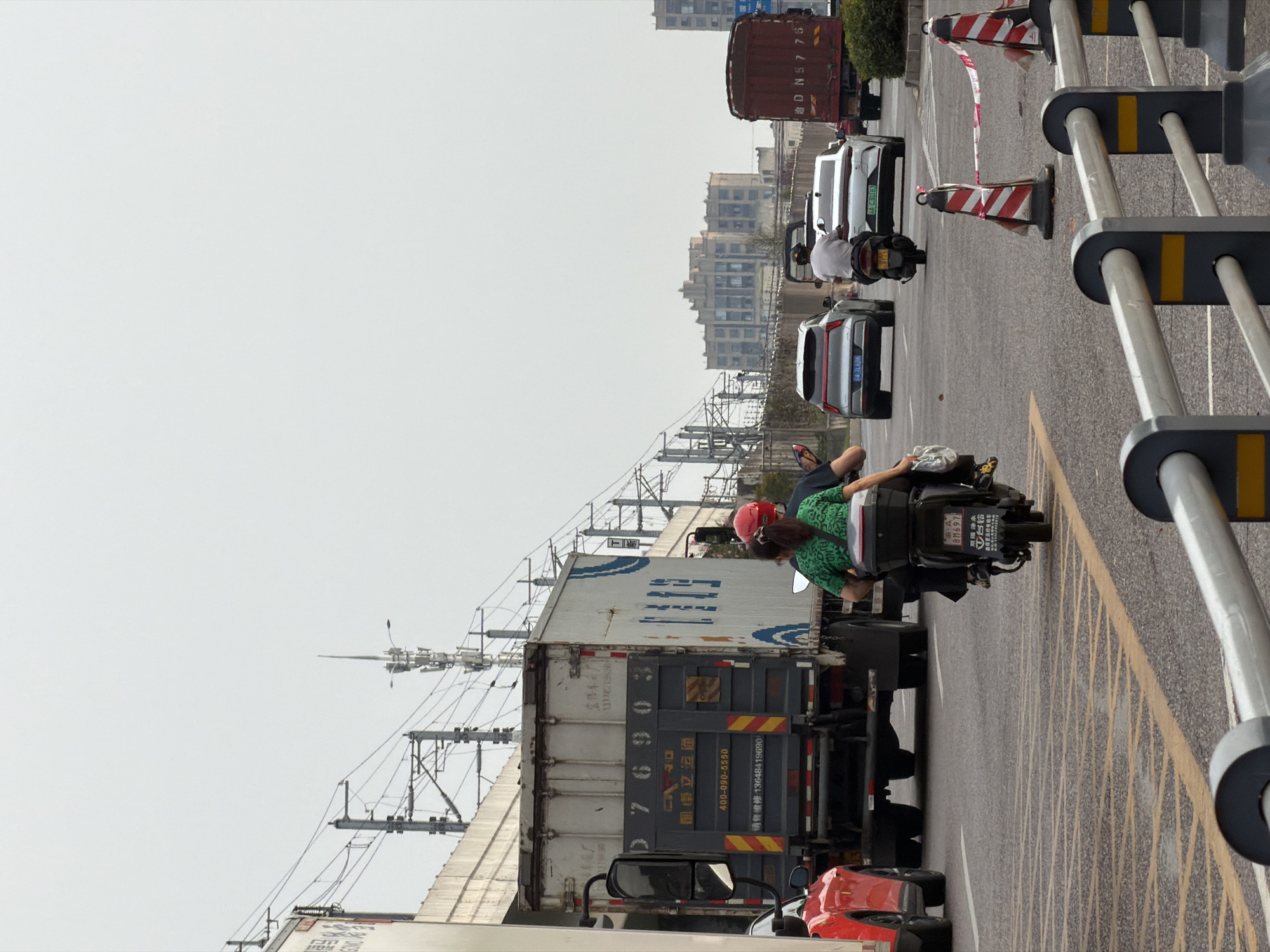 The image captures a traffic scene in Jiangjin, China, likely on a cloudy day. In the foreground, a motorcycle with two riders is moving away from the camera, with the passenger wearing a green top and a red helmet. The motorcycle is followed by a white car. To the left, a red car is partially visible, and a large white and gray semi-trailer truck is stopped. The truck has various markings, including "CARGO," a phone number "400-090-5550," and Chinese characters. Overhead, elevated railway tracks with power lines are visible, supported by concrete structures. Further in the background, a red flatbed truck with a license plate "D N57 75" is in motion. Other vehicles, including silver cars and more distant buildings, are also present. The road surface shows yellow road markings and what appear to be temporary traffic cones with caution tape. The overall environment suggests an urban or suburban area with active transportation infrastructure.