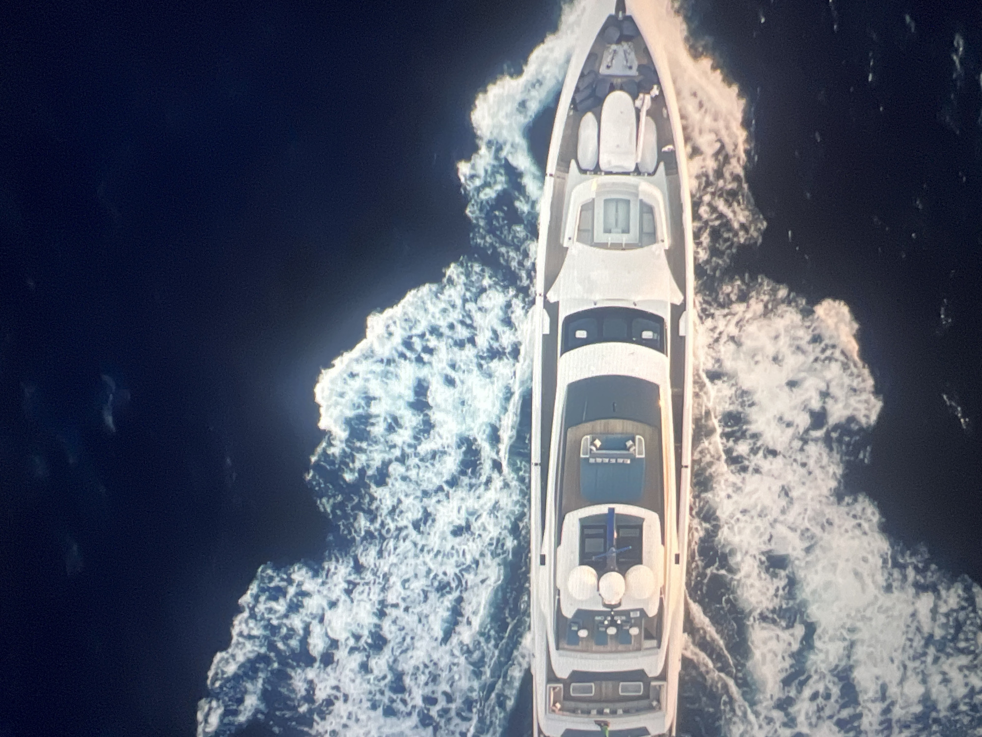 An aerial view captures a long, white yacht cruising through dark blue water, leaving a foamy white wake. The yacht has multiple decks, a helipad at the stern, and various seating areas and structures visible. The water is dark, suggesting deep ocean or night time. The scene is devoid of any urban environment, human figures, or distinguishable geographical features.