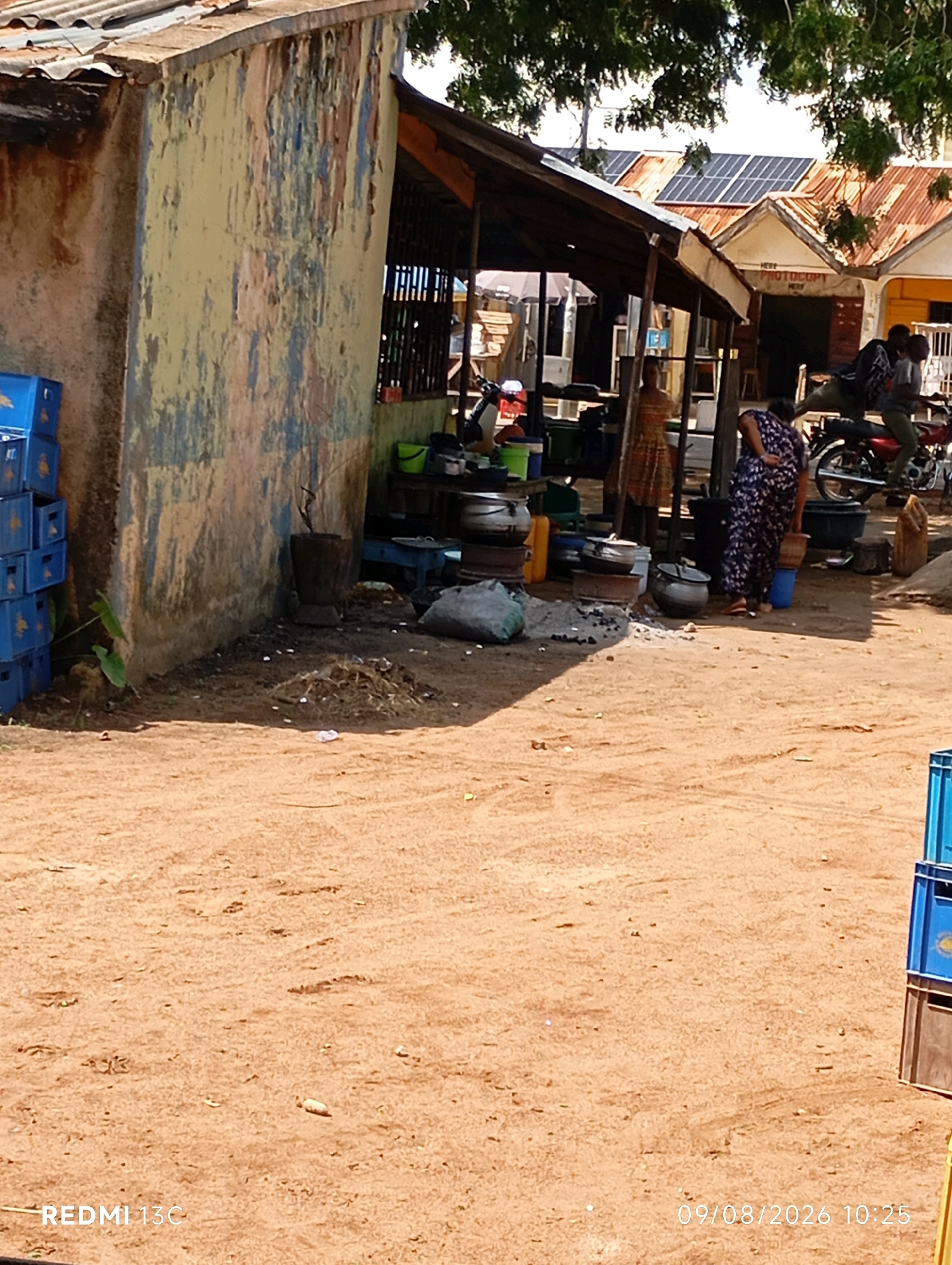 An outdoor scene in Shendam, Nigeria features a rustic marketplace with a weathered building on the left. Blue plastic crates are stacked against the building. A covered stall area extends from the building, displaying various cooking pots and utensils. A woman in a patterned dress is visible in the foreground, holding a pot and standing near a blue bucket. Behind her, a man is seated on a motorcycle. In the background, buildings with corrugated metal roofs, some fitted with solar panels, are visible, suggesting a semi-urban or peri-urban environment. The ground is composed of reddish-brown soil.
