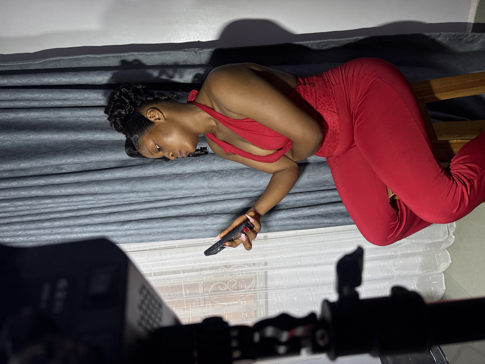 A woman in a red jumpsuit is lying on her side, holding a black remote control in her right hand. Her head is resting on a grey, textured surface, and her body is angled on a wooden surface. She has her eyes closed and appears to be relaxed. Her hair is styled in braids. In the foreground, a black object with a lens is partially visible, suggesting that the photo was taken with a camera. The setting appears to be indoors, with soft lighting casting shadows on the wall behind the woman. The overall mood is tranquil and intimate. There is no visible text in the image. The location context provided is Gura Top, Nigeria.