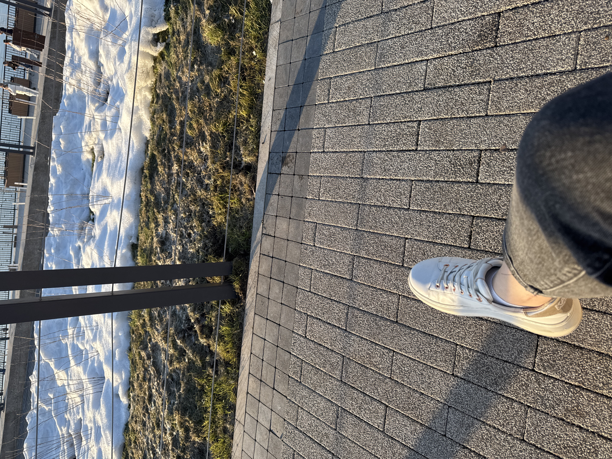 The image shows a view from a paved walkway with a single person's foot and leg in the lower right corner, wearing a white sneaker and dark-colored jeans. The walkway is made of gray interlocking pavers, with a darker gray border running along the edge.
To the left of the walkway is a grassy area partially covered with snow. A black metal fence runs along the edge separating the walkway from the grassy area. In the background, visible behind the fence, are benches and a structure possibly located in a park or recreational area.
The lighting suggests it is daytime with the sun low in the sky, casting shadows across the pavement.
Based on the metadata, the location is Perm, Russia.
