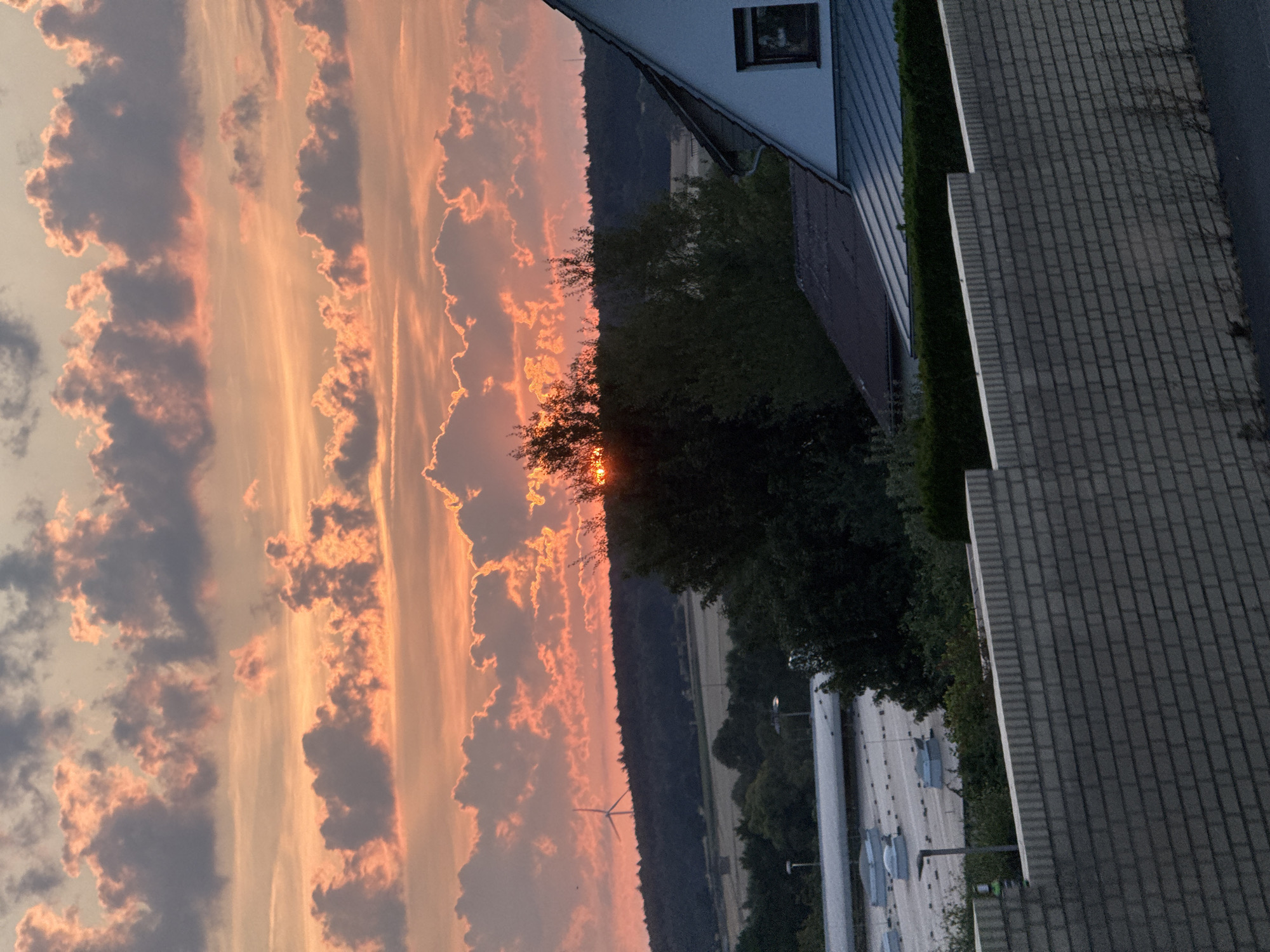 This image captures a sunset over a rural outskirts area of Alsfeld, Germany. The sky is filled with dramatic clouds tinged with orange and pink hues, illuminated by the setting sun which is partially obscured by the clouds and trees. A single wind turbine is visible on a distant, dark treeline. In the foreground, a white brick wall with a textured surface runs horizontally across the lower part of the frame. Beyond the wall, the roof of a building with skylights is visible, and further in the background, part of a white house with a dark roof and a single window is seen. The scene appears to be a quiet, residential or semi-industrial area at dusk.