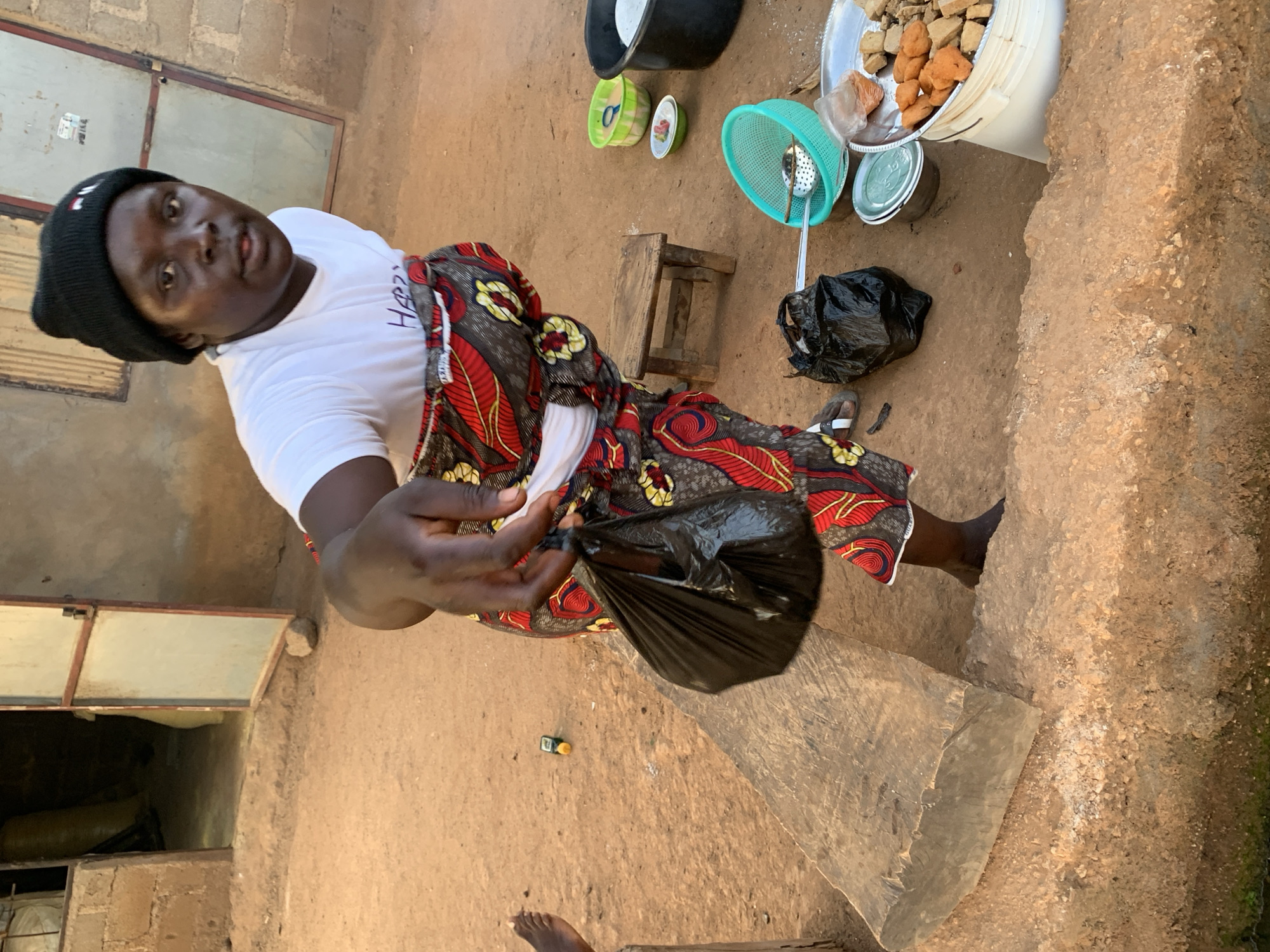 A woman wearing a black beanie, white T-shirt, and patterned fabric is holding a black plastic bag in Agban, Nigeria. She stands in a sandy area near a building with an opening. Behind her are cooking utensils, including bowls, a colander, and a bucket containing fried dough.