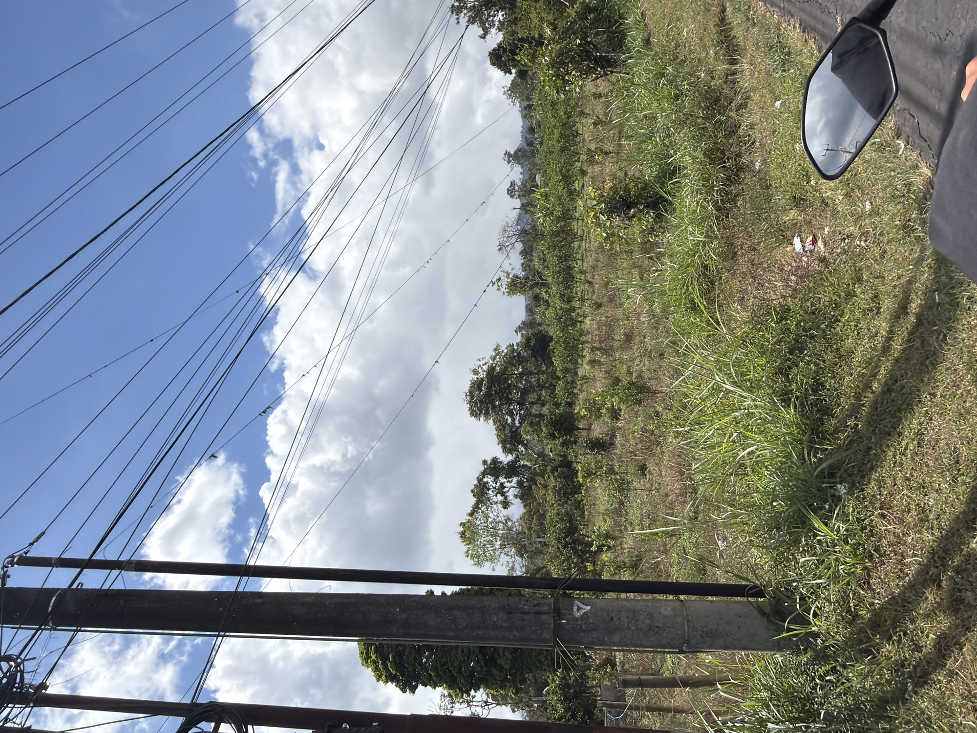 The image depicts a roadside view in Kintamani, Indonesia, characterized by a landscape dotted with vegetation and a network of overhead power lines. In the foreground, a concrete utility pole stands to the left, with a motorcycle's side mirror visible on the right, reflecting the sky and utility lines. Beyond the pole, a field stretches out, sparsely covered with dry grass and small saplings, interspersed with taller trees in the middle ground. The background reveals a denser arrangement of trees, with a hint of mountainous terrain beneath a partly cloudy sky. The overall impression is one of a rural or semi-rural setting with evidence of infrastructure and cultivated land.