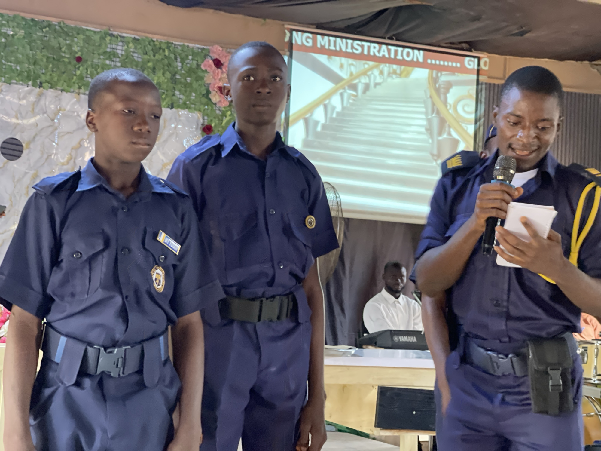 An indoor scene in Akwanga, Nigeria, features three young men in matching navy blue uniform shirts and trousers, along with a man in a white shirt in the background. The uniform shirts have pockets and button closures, with shoulder epaulets visible on the speaker and the middle figure. The leftmost individual has a nametag reading "MY YUSUF" and a gold emblem on his left chest pocket. The middle individual also has a similar gold emblem. All three young men wear black belts with plastic buckles; the speaker also has a black utility pouch attached to his belt.
On the right, a man with yellow epaulets is speaking into a black microphone, holding a small white notepad. He appears to be smiling. To his left, the middle man stands facing forward, looking attentive. The man on the far left stands looking slightly to his right, also appearing attentive.
Behind them, a projector screen displays an image of a grand, ornate staircase with golden railings and steps, beneath text that reads "NG MINISTRATION" and "GLC" (partially visible). The backdrop behind the screen features a white textured material adorned with green leafy garlands and pink flowers.
In the background, positioned behind a wooden table or platform, another man in a white collared shirt is visible, seated behind a "YAMAHA" brand keyboard. A black floor fan is partially visible at the bottom right. The setting suggests a formal event, possibly a religious service, presentation, or ceremony within a church or community hall.