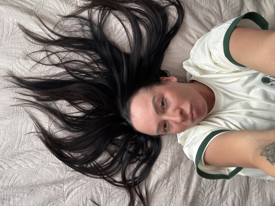 A single adult female subject is depicted lying on her back on a light beige, quilted surface with a repeating leaf-like pattern. Her long, dark brown to black hair is fanned out extensively above her head. The subject's face is directed towards the camera, exhibiting a slight pout with pursed lips. She is wearing a cream-colored t-shirt with dark green banding on the short sleeves; a small circular logo featuring a tennis racket and ball is present on the left chest. A dark tattoo, resembling an insect wing, is visible on her left forearm. The image appears to be a self-portrait taken indoors under diffuse lighting conditions. The location is the city of Pavlovsky, Russia.
