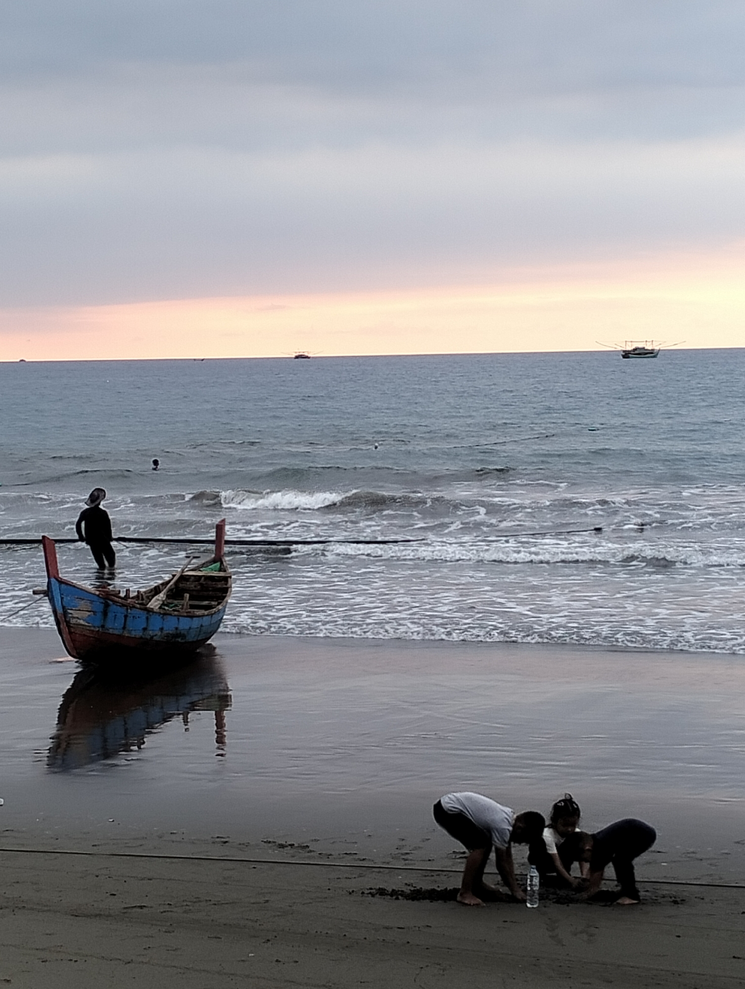 The image depicts a beach scene in Uleekareung, Indonesia, likely during twilight given the soft lighting and muted colors. In the foreground, three children are playing in the sand, two appearing to be digging or building. A bottle of water is visible near them. In the mid-ground, a weathered, colorful wooden fishing boat is partially submerged in the shallow water at the shoreline. A single person, dressed in dark clothing, stands in the water near the boat, facing away from the camera. The ocean extends to the horizon, with two small fishing boats visible in the distance. The sky is overcast with hues of pink and orange suggesting a sunset or sunrise. The wet sand reflects the sky and the boat.