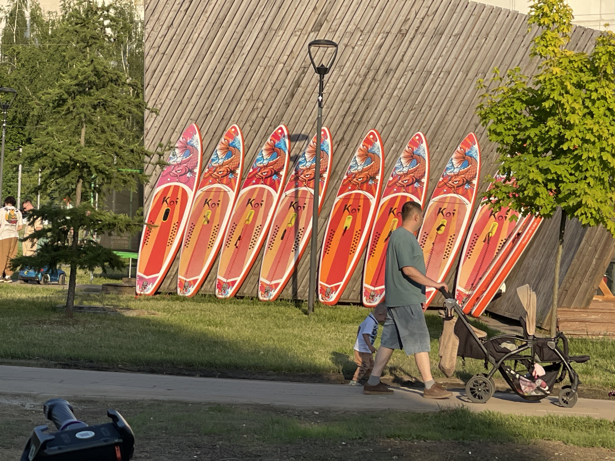 The image displays an outdoor scene in Moscow, Russia, featuring a paved pedestrian path, green grass, and a tall, diagonally slatted wooden wall as a background element. Nine colorful stand-up paddleboards (SUPs) are propped vertically against the wooden wall, each predominantly red and orange with distinct graphic designs of koi fish in various colors (blue, pink, and orange). The brand name "Koi" is visible on multiple boards. A dark grey lamppost with a modern fixture stands centrally in front of the wall and SUPs.
On the paved path in the foreground, a man, wearing a dark green t-shirt, light grey shorts, and brown shoes, walks towards the right while pushing a black double stroller. A small child, dressed in a light-colored t-shirt and light brown pants, walks beside the man, positioned to his left. A brown garment hangs from the stroller's handlebar, and items are visible in the stroller's lower storage basket.
In the mid-ground on the left, a small coniferous tree stands, with a blue child's electric toy car visible on the grass behind it. Further back on the left, several indistinct figures are present near other trees. On the far right, a bright green deciduous tree partially obscures the last two paddleboards. In the bottom left corner, a black remote control device rests on the grass. The scene is illuminated by natural light from the right, casting shadows towards the left.