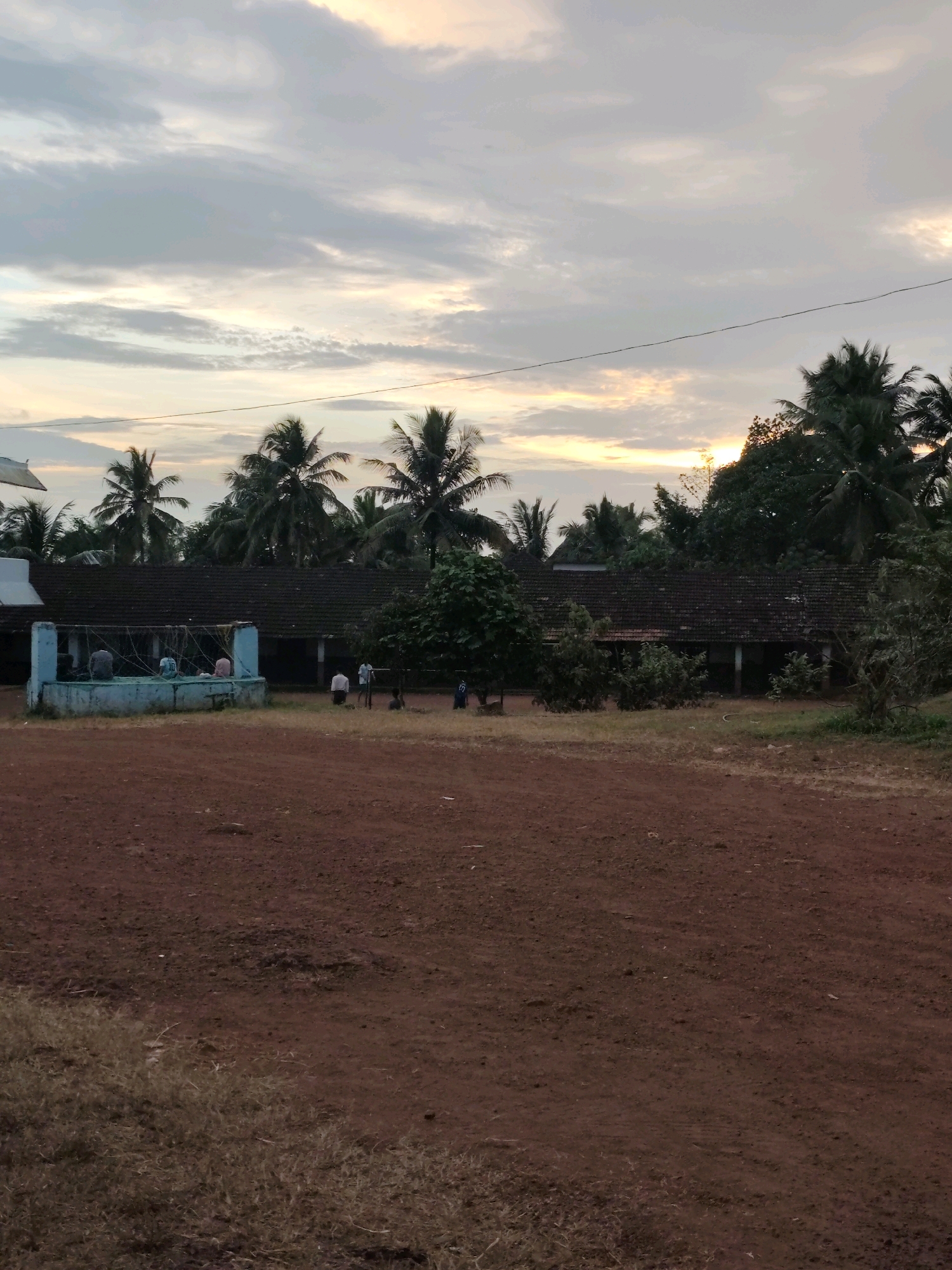 The image depicts a school or community building in Kuttippuram, India, during what appears to be late afternoon or early evening, with a dusky sky and diffused sunlight. In the foreground, a reddish-brown dirt area is visible, with some sparse dry grass on the left. In the mid-ground, a long, tiled-roof building runs horizontally across the frame. To the left of the building, there is a small, light blue structure with a railing. Within this blue structure, several individuals are seated, suggesting a communal gathering or activity. Further to the right, along the front of the main building, a few individuals are standing or engaged in activities. Behind the building and to the sides, a dense growth of palm trees and other vegetation creates a lush backdrop. The overall impression is one of a rural or semi-rural setting.
