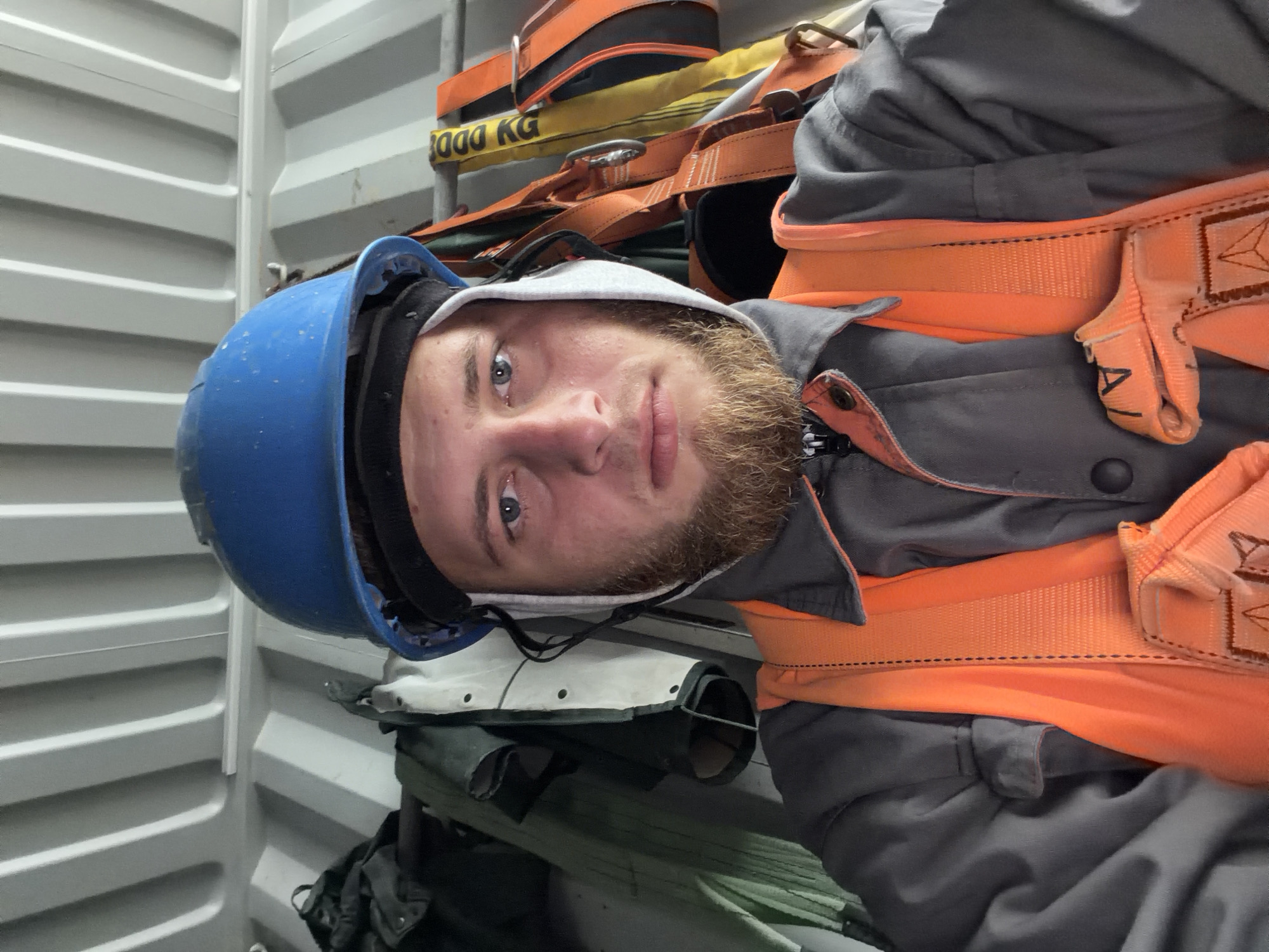 A close-up, front-facing selfie shows a person wearing safety gear. The person has fair skin, blue eyes, and a light brown beard and mustache. They are wearing a blue hard hat, an orange safety vest over a gray work jacket, and what appears to be a grey hood. The background consists of corrugated metal walls and ceiling, with various pieces of work equipment, including harnesses, hanging on a rack. The lighting is bright and direct, casting shadows. The image quality is good, with sharp focus on the subject. The location is not specified in the image.