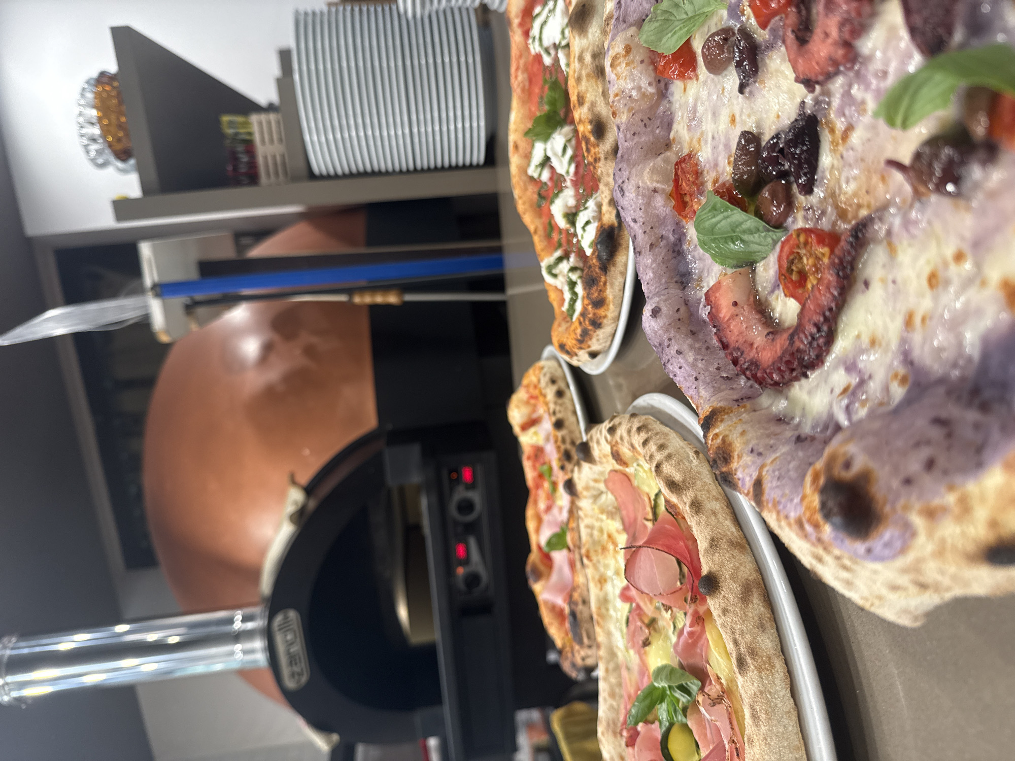 This image captures a close-up view of several pizzas displayed in front of a traditional wood-fired oven, likely in a restaurant in Trento, Italy. The oven, a prominent feature, is large and dome-shaped, with a visible opening and a metallic chimney. To the right of the oven, a gray shelving unit holds a stack of white plates, some yellow boxes, and a decorative, amber-colored object.
In the foreground, multiple pizzas are arranged on white plates. One pizza is topped with prosciutto, zucchini, and basil. Another appears to have a red sauce base with mozzarella and basil. The closest pizza, partially visible, has a distinctive purple-tinted crust and is topped with octopus tentacles, cherry tomatoes, olives, and basil. The pizzas show signs of being freshly baked, with charred crusts and melted cheese.
The overall environment suggests a culinary setting, with the focus on the pizzas and the pizza-making equipment. The lighting is warm, highlighting the textures and colors of the food. There are no people visible in the image.