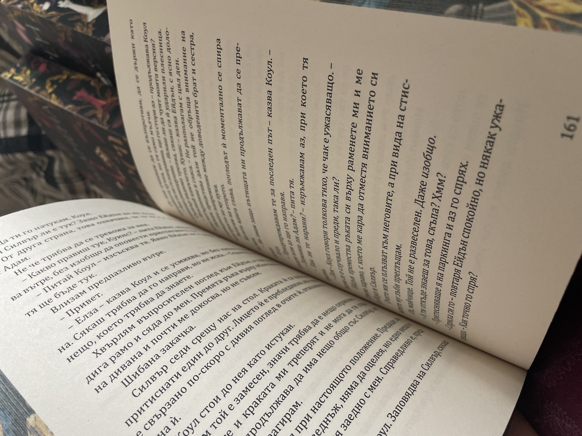 Here is a detailed description of the image:
**Scene:** The image depicts an open book with text in Bulgarian, laying on what appears to be a burgundy or dark pink surface, possibly a bedspread. The book is open to two pages, with the right page more visible. The text is closely spaced and formatted as dialogue within a narrative. The page number '161' is visible in the bottom right corner of the right page.
**People:** Although there are no people visible in the image, the text indicates a narrative involving characters named Коул (Koul), Ейдън (Aiden), Силвър (Silver), Куинс (Quins), Елза (Elza), and Адам (Adam). The dialogues imply relationships and interactions between these characters.
**Subjects:** The subject of the image is the text from a Bulgarian book. The text details an interaction between characters in a narrative.
**Objects:** The primary object is the book itself. Other objects mentioned in the text include a стол (chair), дивана (divan), and паркинга (parking).
**Actions:** Actions mentioned in the text include talking, stopping, looking, smiling, sitting, trembling.
**Interactions:** The text describes a tense exchange between characters, indicating conflict and potential threats. There is talk of deception, pain, and a foreboding sense of danger.
**Environment:** The environment is ambiguous, but the mentions of дивана (divan) and паркинга (parking) imply indoor and outdoor settings, respectively.
**Location:** There is no information provided about the location in the city of Zlatograd, Bulgaria.