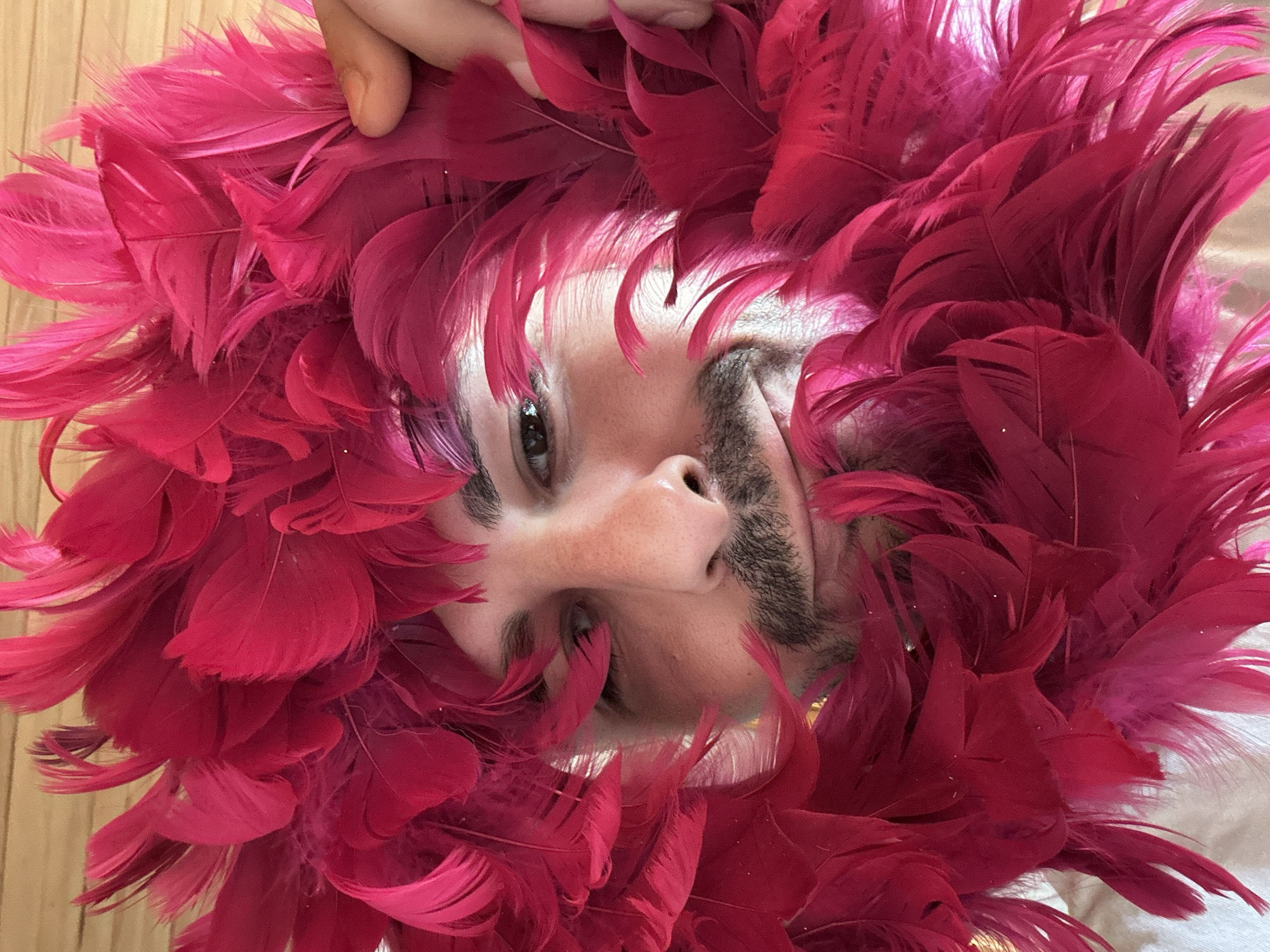 A medium shot presents a person from a low angle, surrounded by a vibrant array of pink feathers. The person's face is centered, with eyes looking directly at the camera. Visible facial features include a mustache and beard. A fair-skinned hand is present at the top of the frame, partially obscured by feathers. The background is out of focus, showing a wooden floor. There is no indication this picture was taken in Slavyani, Bulgaria.