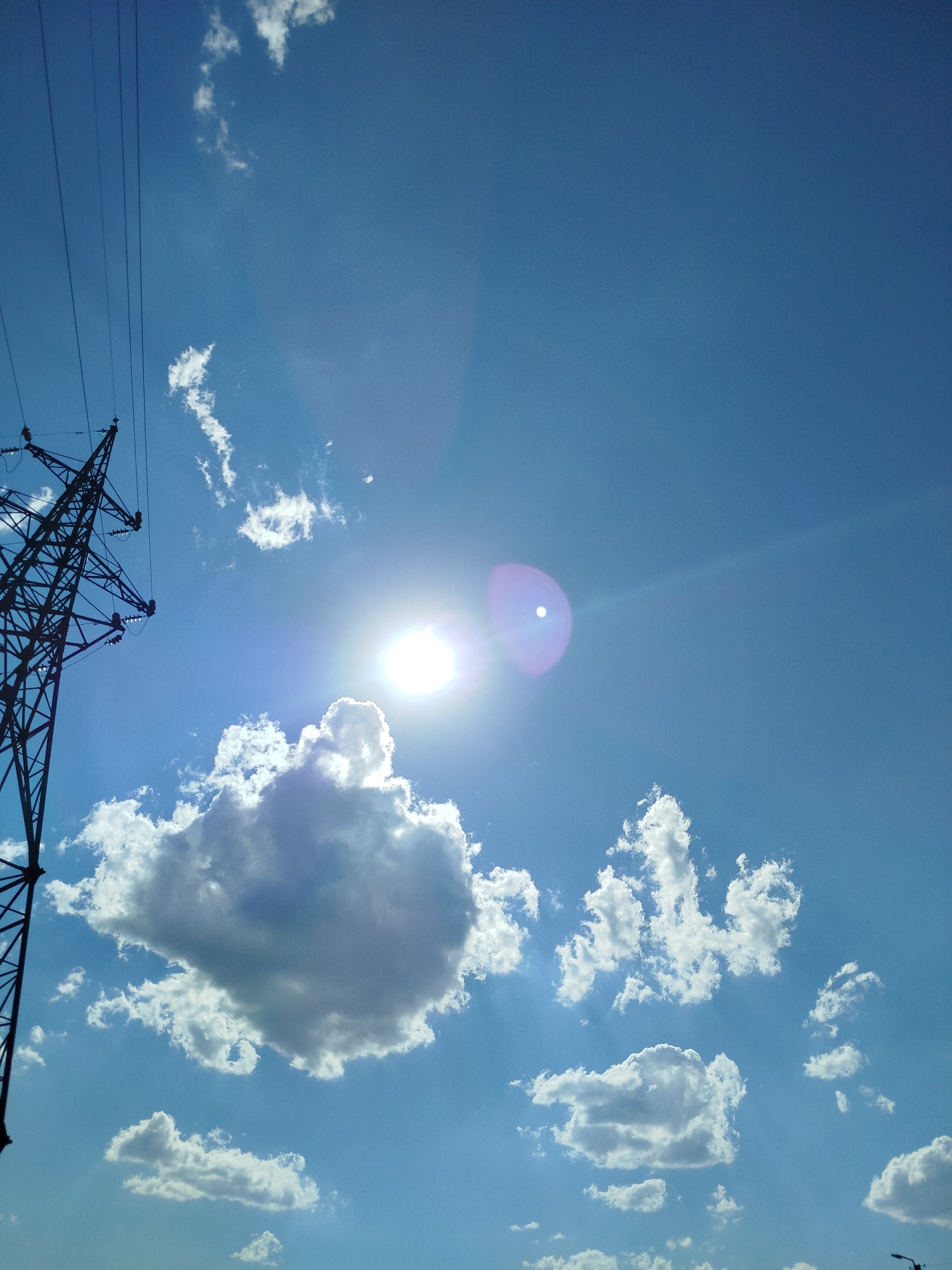 The image shows a view of the sky with a large electrical transmission tower on the left. The tower is dark and silhouetted against the bright blue sky. Several white, fluffy clouds are scattered across the sky, with the sun prominently visible in the upper center, emitting a bright glare. There are no people or specific landmarks visible that would indicate the exact location within Qarağandı city administration, Kazakhstan. The environment is an outdoor setting, likely on a clear day with some cloud cover.