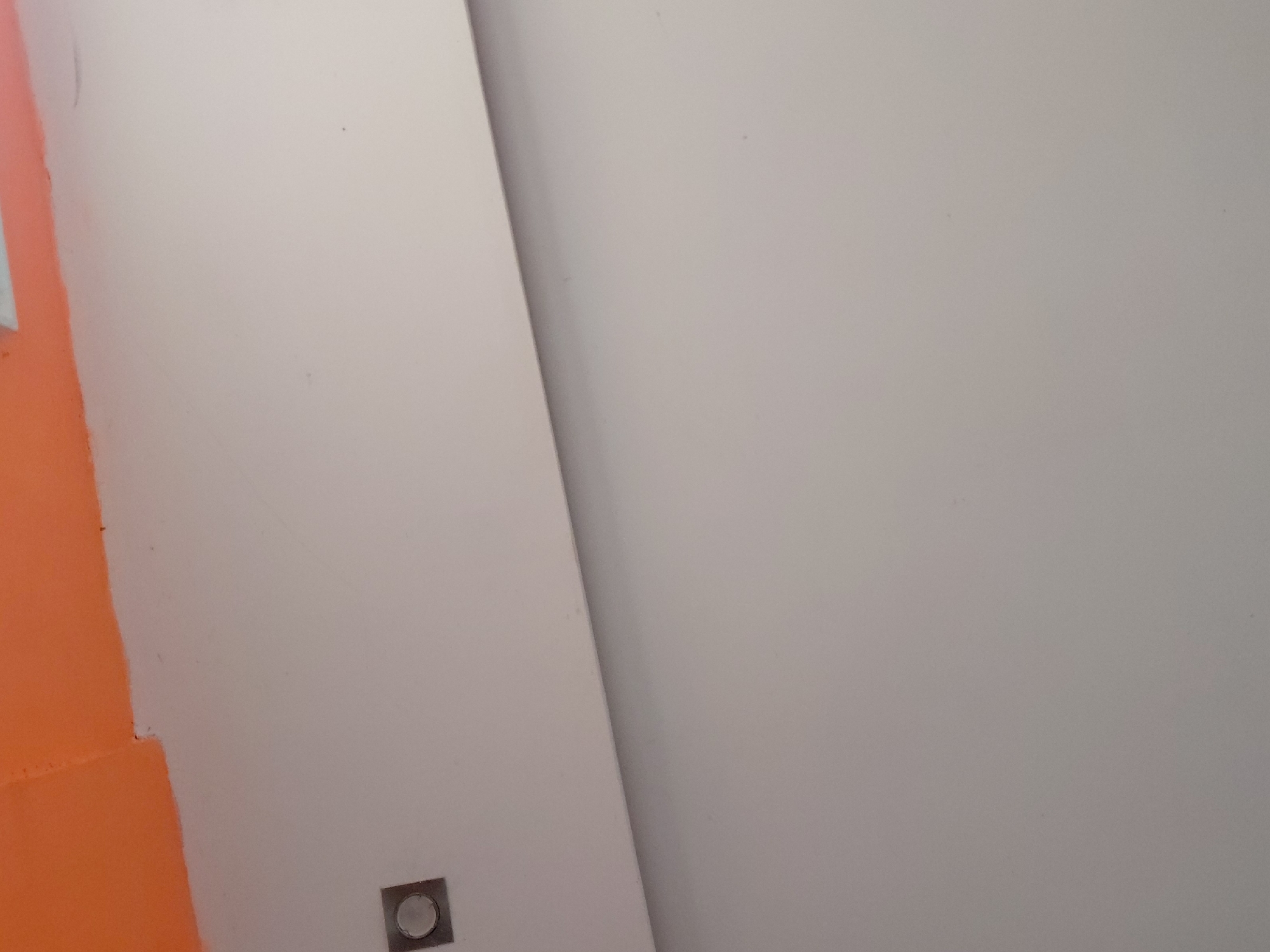 The image displays an abstract, close-up view of an interior architectural space, predominantly featuring two large, contrasting color blocks separated by a diagonal line. On the left, approximately one-quarter of the frame is taken up by a vibrant orange surface, likely a painted wall or partition, which shows a slightly textured finish. The central and right portions of the image are dominated by a smooth, light gray or off-white surface, also appearing to be a wall or panel.
A sharp, dark diagonal line extends from the top-center to the bottom-right, clearly demarcating the boundary or joint between these two main surfaces, suggesting a corner, a door frame, or a structural division. In the lower-left corner of the light gray surface, near the orange section, a small, square metallic fixture is visible. This fixture has a circular indentation or button in its center, resembling a switch, an intercom button, or a similar electrical component.
No people are present, and no activity is occurring. The lighting appears even, suggesting an indoor setting with no specific time of day discernible. There are no discernible location cues within the image itself to confirm the provided context of Santa Ana, Venezuela. No visible text is present.