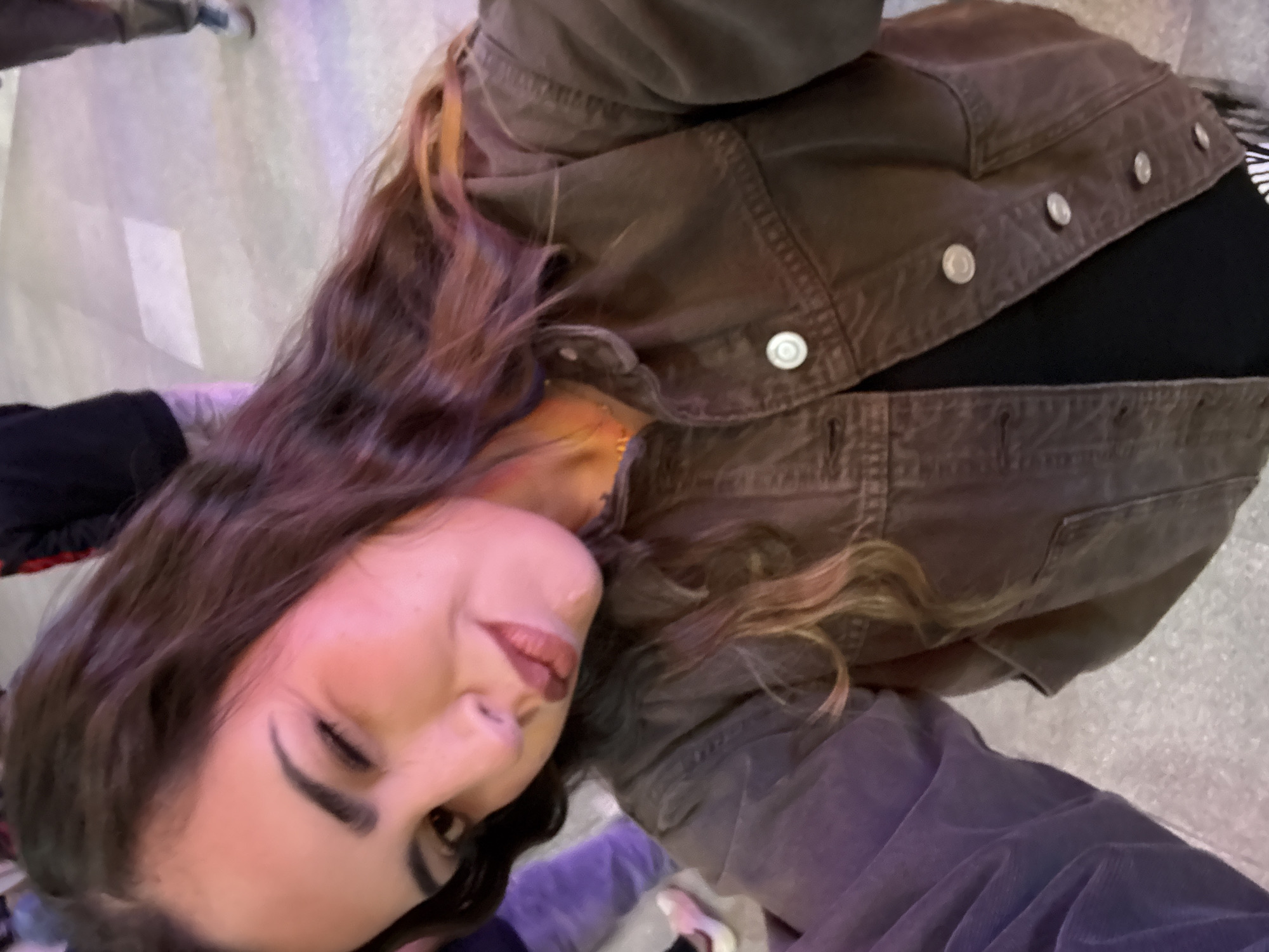A woman is seen in a selfie shot. She has long brown hair and is wearing a brown denim jacket over a black top. The background appears to be an indoor location with a tiled floor and other people partially visible. It is unclear from the image if the location is in the city of Puebla, Mexico.