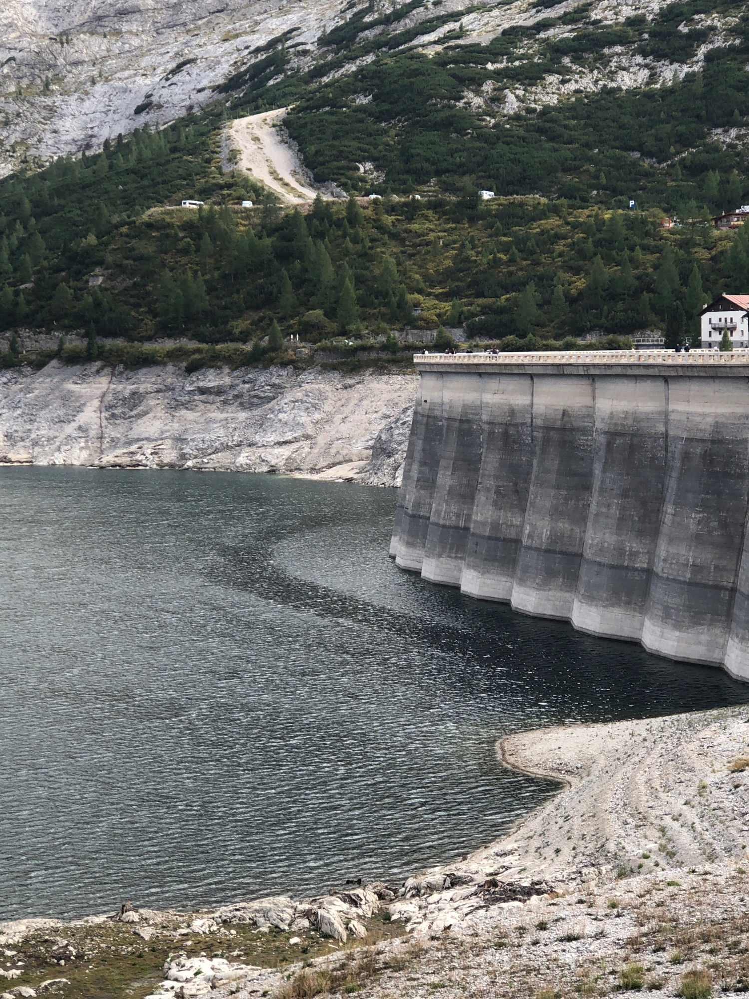The image shows a large concrete dam with a body of dark water in the foreground. The dam wall is curved and has vertical segments. There is a paved walkway on top of the dam, with several people visible walking along it. To the right of the dam, a white building with a red roof is situated, and further back, the slopes of a mountain are covered with dense green trees and lighter patches of bare rock. A dirt road winds up the mountainside, with a few white vehicles parked along it. The water's edge meets a rocky, sparsely vegetated shoreline. The scene appears to be a mountain reservoir. The dam is likely the artificial lake of the Fedaia Dam. Canazei, Italy, is in the Fedaia Valley, a location known for its mountain landscapes and dams, making this a plausible scene from that region.