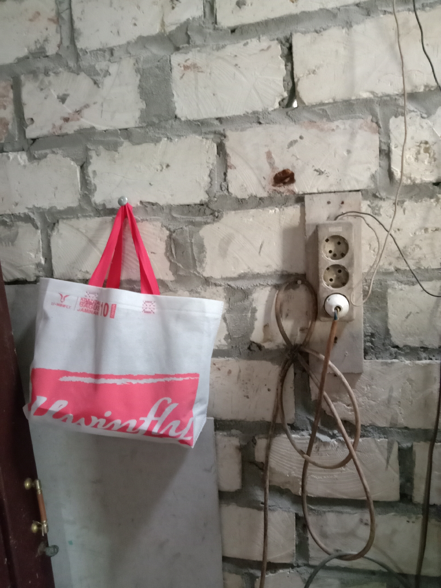 In an indoor scene, likely a workshop or utility area, a white fabric tote bag with pink handles hangs from a silver hook on a white brick wall. The bag displays pink lettering that appears to say "Levis." To the right of the bag, a weathered electrical outlet box is mounted on the wall, containing two circular sockets and a single white plug with a brown cord. Various electrical wires are visible, some coiled and others extending out of frame. The wall itself is constructed of rough, off-white bricks with visible mortar lines. The floor appears to be a flat, light-colored surface. The image is taken from a medium distance, with the focus on the bag and the electrical outlet. There is no indication of the specific location within Jumput, Indonesia.
