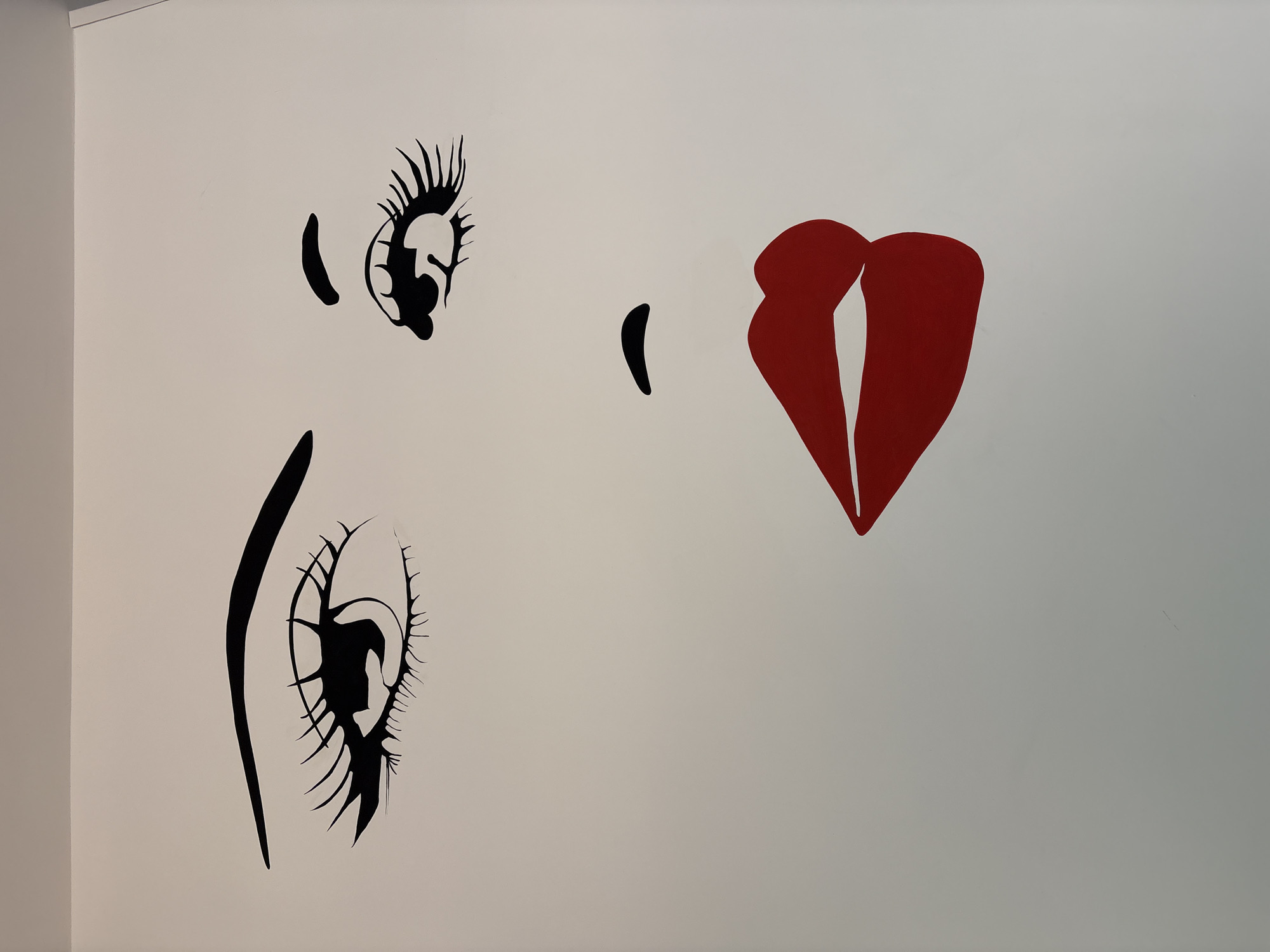The image depicts a section of a white wall with a minimalist artistic representation of a face. The features are stylized and graphic. Two eyes are drawn in black, with prominent eyelashes. To the right of the eyes is a single, red, abstracted representation of lips. The setting is indoors, likely a room in a building. This artwork is in Rome, Italy.