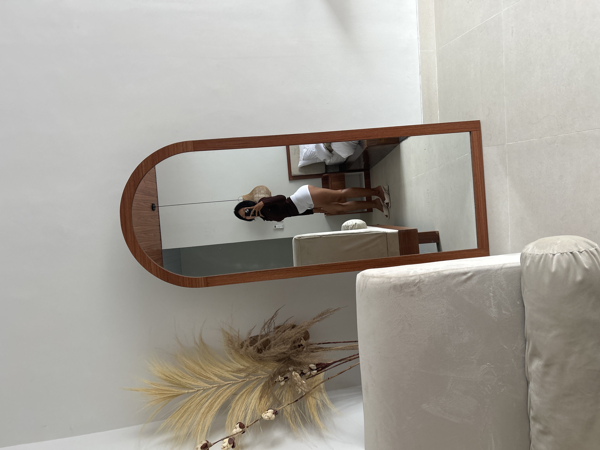 A full-length, arched wooden mirror stands on a light-tiled floor against a white wall. A woman is visible in the mirror, taking a selfie. She is wearing a dark-colored top and white shorts, with light-colored heeled mules. To the left of the mirror, a large, pale upholstered couch is partially visible. Behind the couch and to the left of the mirror, a bouquet of dried pampas grass and cotton branches is arranged. The reflection in the mirror also shows a bed with white linens and a bedside table, suggesting the room is a bedroom. The lighting appears to be natural, coming from an unseen window. The environment suggests a modern, minimalist interior design. The location is specified as Abiansemal, Indonesia.