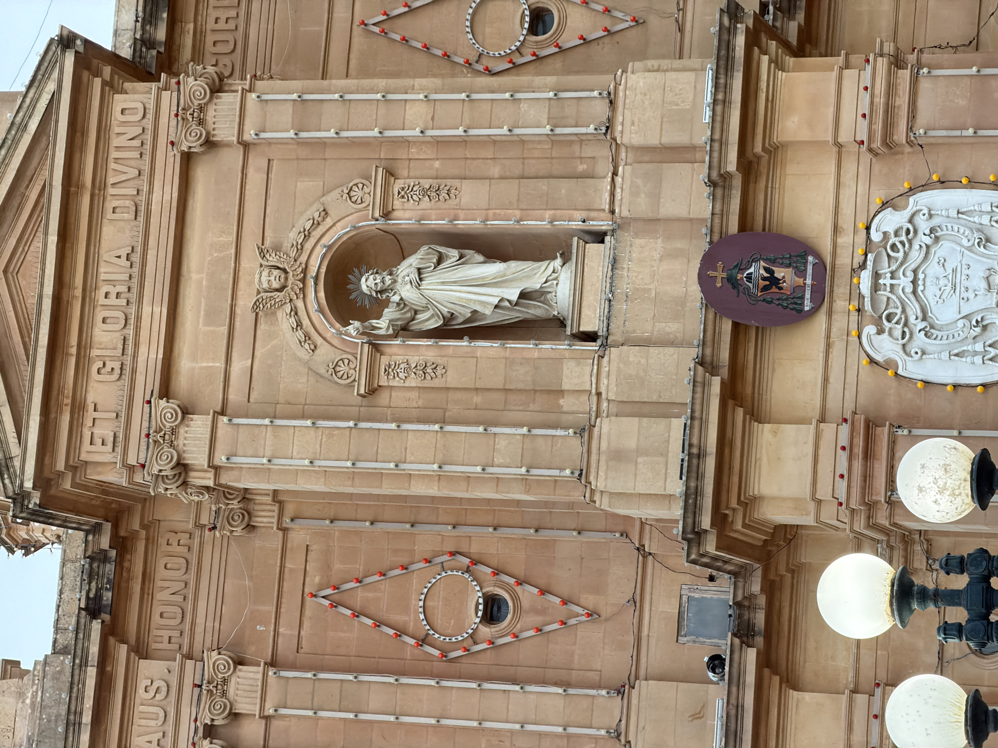 This is a detailed, eye-level, medium shot of the facade of the National Shrine of the Sacred Heart of Jesus in Fontana, Gozo, Malta. The facade is constructed of warm-toned stone blocks. The upper portion of the building features the inscription "AUS. HONOR ET GLORIA. DIVINO." above a central niche containing a statue of Jesus Christ. The statue depicts Jesus with his right hand raised and his left hand on his chest, wearing a flowing robe and a halo. Below the niche is an oval coat of arms featuring a black eagle clutching a cross. The building is adorned with decorative elements, including ornate columns and diamond-shaped patterns with red lights. In the foreground, three large, spherical, illuminated lamps are visible. The sky above is overcast.