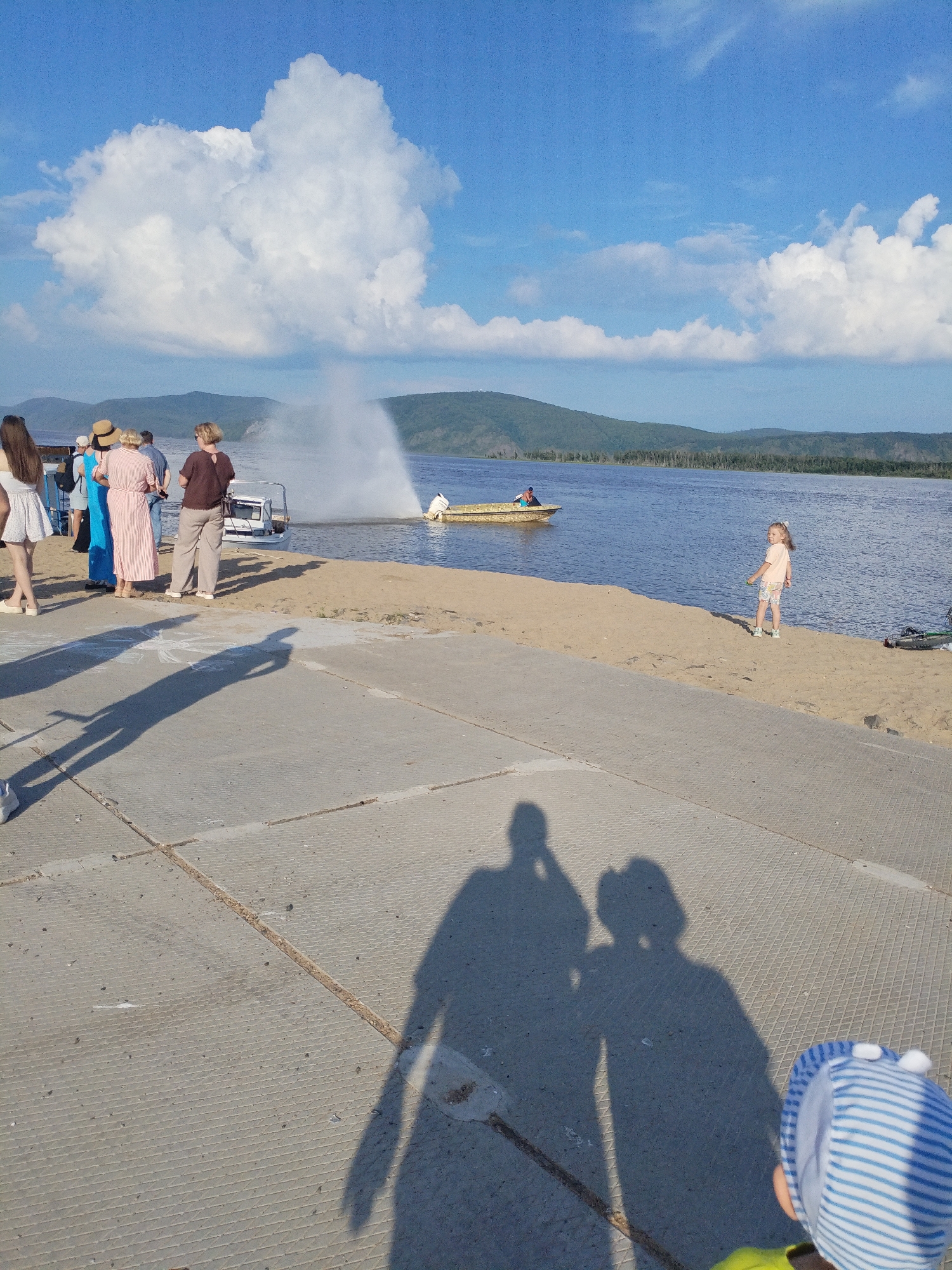 The image depicts a scene on the bank of the Amur River in Komsomolsk-on-Amur, Russia. Several individuals are visible on a sandy embankment and a concrete walkway. In the foreground, the shadows of people are cast on the concrete. To the left, a group of people stands, some facing the water. One person is wearing a white dress, another a blue dress, and a third a striped dress. Further back, a small boat is anchored near the shore, with a person standing on it. In the middle ground, a child with blonde hair is standing on the sand, looking towards the viewer. The Amur River is calm, with hills and trees forming the distant shoreline. The sky is blue with scattered white clouds. There is a jet of water spraying from the river, likely from a boat's engine or a water feature.