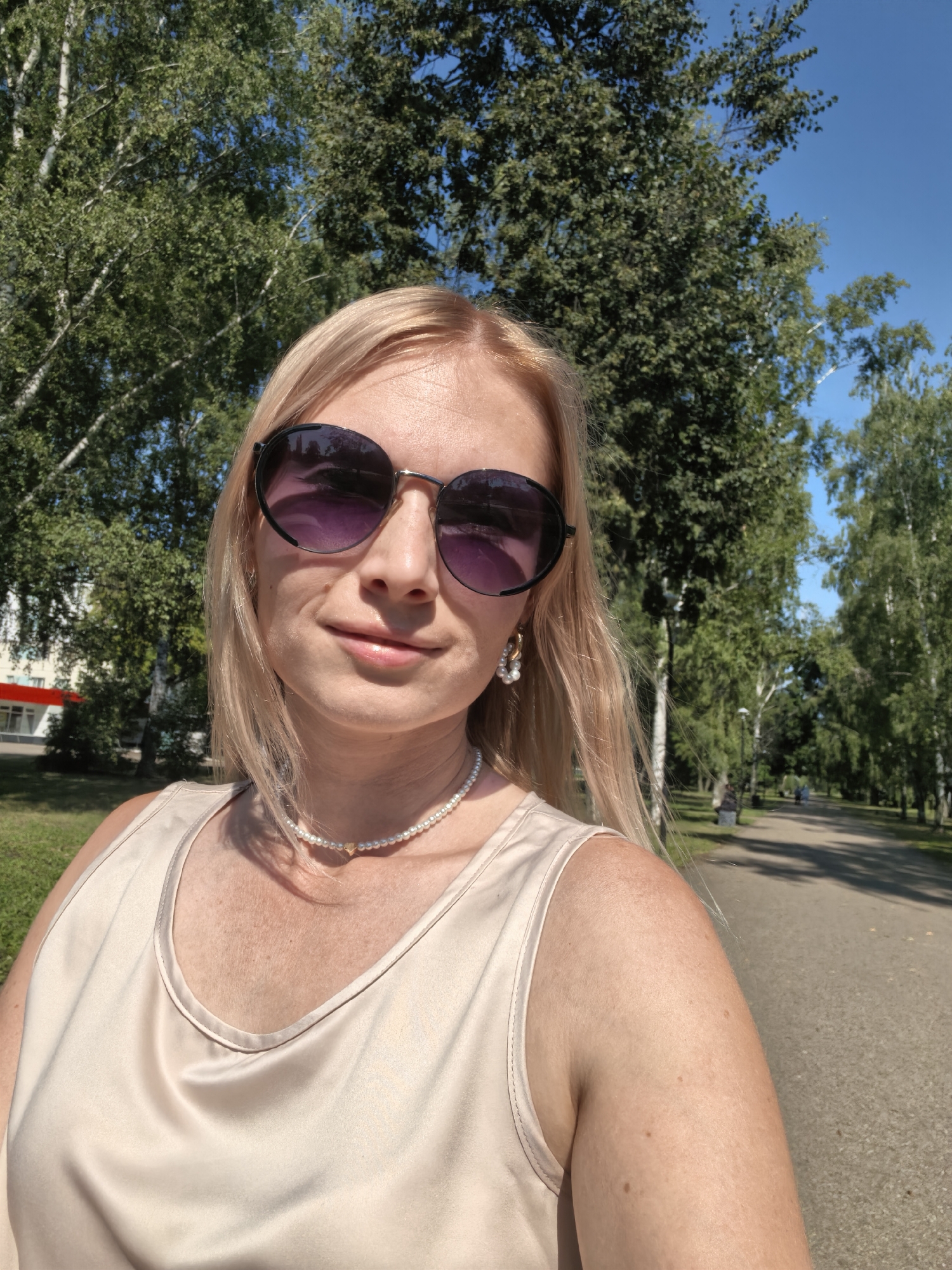 This is a selfie taken outdoors in Sterlitamak, Russia. The subject is a fair-skinned woman with blonde hair, wearing large, round, dark-lensed sunglasses, a pearl necklace with a heart-shaped pendant, and a light-colored, sleeveless top. She is positioned in the foreground, with a park-like setting behind her. Trees line a paved pathway, where a few other people can be seen walking in the distance. The sky is clear and blue, indicating a sunny day. A building with a red roof is visible to the left of the frame, suggesting an urban park environment.